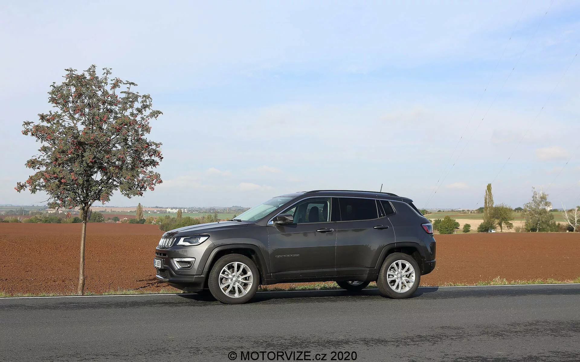 Imagem de um SUV Jeep Compass 2017 estacionado em uma estrada de asfalto plana ao lado de uma jovem árvore com folhas sob um céu claro e diurno. O ângulo lateral captura o perfil do veículo de para-choque a para-choque, destacando seus elementos de design moderno, como a grade frontal proeminente, faróis elegantes, rodas de liga leve multirraio, espelhos laterais estendidos com possíveis sinais de pisca-pisca e vidros escurecidos. O SUV apresenta um esquema de cores em dois tons e um rack de teto, com reflexos indicando acabamentos cromados na fachada frontal. O fundo rural sugere versatilidade para diferentes terrenos. Marca d'água 'MOTORVIZE.cz 2020'.
