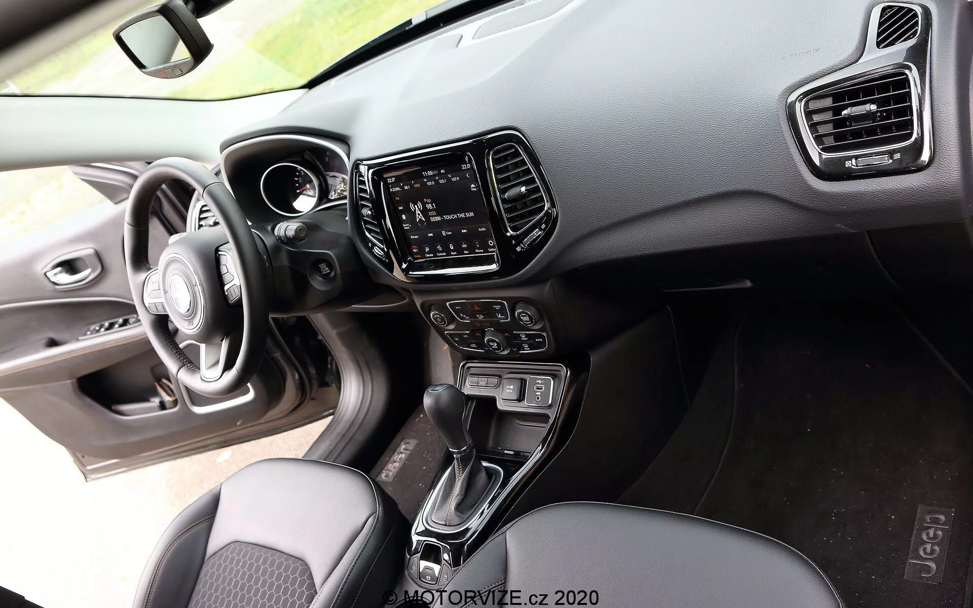 Vista interior de um Jeep Compass 2017 a partir da perspectiva do passageiro, exibindo um volante de três raios com botões de controle integrados, um painel de instrumentos digital parcialmente visível no painel feito de materiais macios ao toque e plástico duro com costura visível, um sistema de infoentretenimento com tela sensível ao toque no console central ladeado por saídas de ar verticais, uma alavanca de câmbio automática entre os bancos dianteiros, botões e manípulos para controle de clima, bancos de tecido com design estampado e uma visão do painel da porta do motorista com controles de ajuste de janelas e espelhos laterais realçados por iluminação ambiente, destacando o foco no conforto e tecnologia do segmento de mercado de médio a alto padrão.
