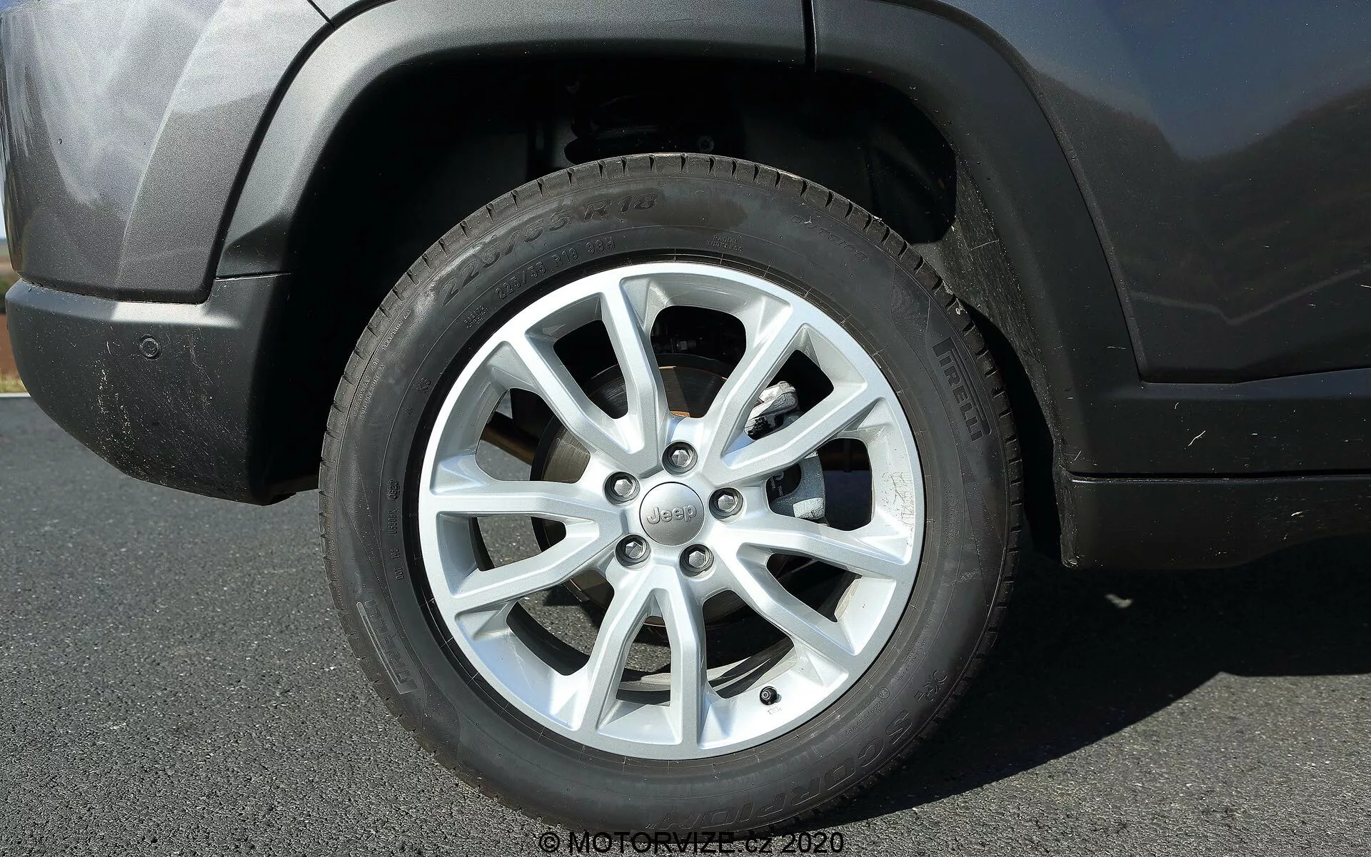 Vista aproximada da roda dianteira do lado do motorista de um Jeep Compass 2017, com um design de liga de 10 pares de raios e acabamento prateado. A tampa central exibe o logotipo da Jeep. A roda e o pneu mostram uma profundidade moderada de banda de rodagem e pequenos sinais de sujeira, indicando uso recente. A foto foi tirada de um ângulo lateral baixo, mostrando parte do arco da roda, do para-choque dianteiro e da superfície asfáltica abaixo. A luz natural e as sombras claras sugerem um dia ensolarado.