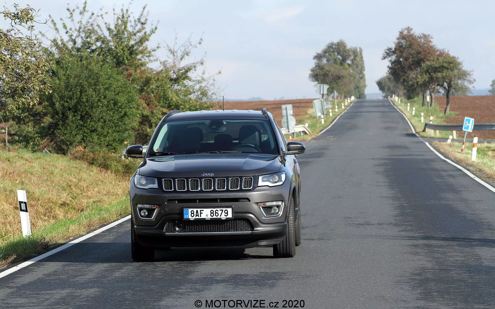 Vista frontal de um Jeep Compass 2017 dirigindo a uma velocidade moderada em uma estrada rural. A imagem captura os detalhes frontais distintos, incluindo a icônica grade frontal de sete fendas, faróis retangulares modernos e luzes de neblina integradas no para-choque. O carro é fotografado ao nível dos olhos, destacando seu design exterior e tecnologia de iluminação sob boa luz natural diurna. Uma placa de licença no estilo europeu está montada centralmente na frente, com uma paisagem rural clara ao fundo.