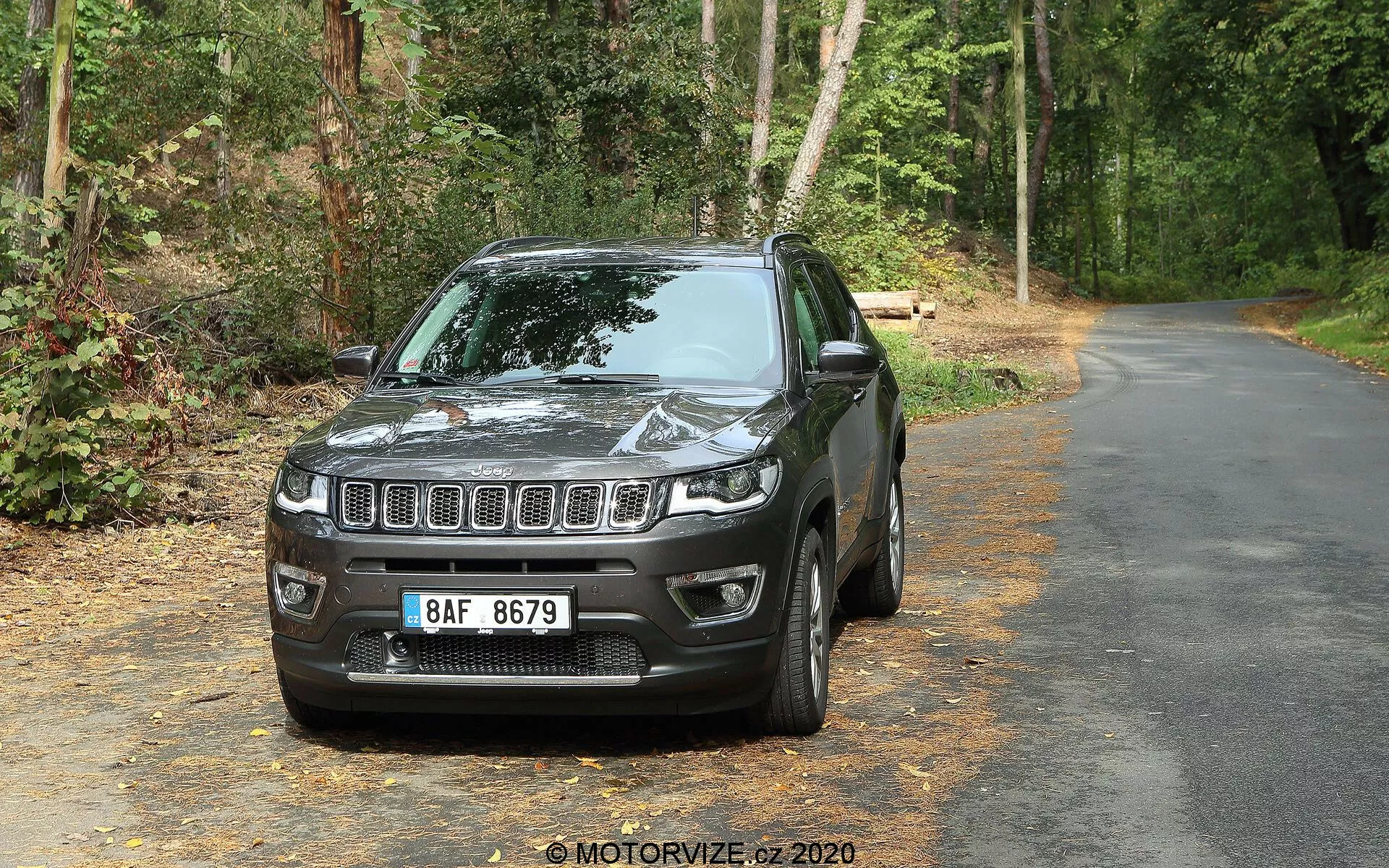 SUV Jeep Compass 2017 em estrada pavimentada cercada por vegetação, de frente para a câmera em um leve ângulo à esquerda mostrando a frente e o lado do motorista. Características visíveis incluem a grade frontal com ripas horizontais com emblema central, faróis retangulares de LED, alojamentos de luzes de neblina no para-choque inferior, capô contornado refletindo luz, capas de espelhos da cor do veículo, janelas levemente escuras, roda de liga de múltiplos raios e para-lamas delineados. O fundo apresenta árvores e folhagem que indicam uma área de estrada arborizada.