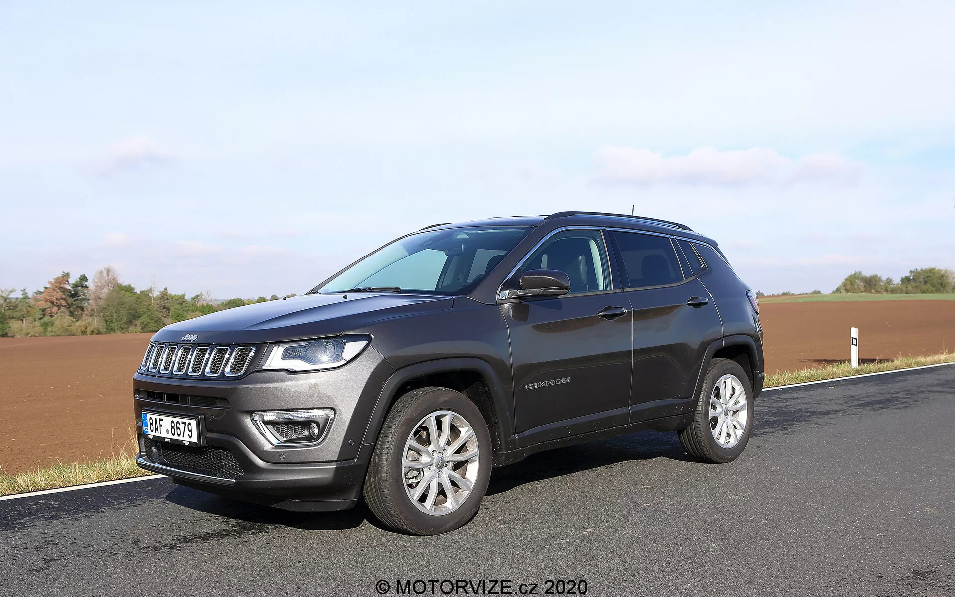 SUV moderno estacionado ao lado de uma estrada com um campo ao fundo, capturado em um ângulo frontal de três quartos. A foto destaca a grade frontal de sete fendas do SUV, os faróis distintos equipados com LED, rodas de liga leve e pintura metálica escura. Detalhes laterais como a linha das janelas, maçanetas das portas, espelhos laterais possivelmente com piscas integrados e uma linha do teto ligeiramente inclinada são visíveis. Um emblema na porta dianteira também é visível, fornecendo informações potenciais sobre o nível de acabamento.
