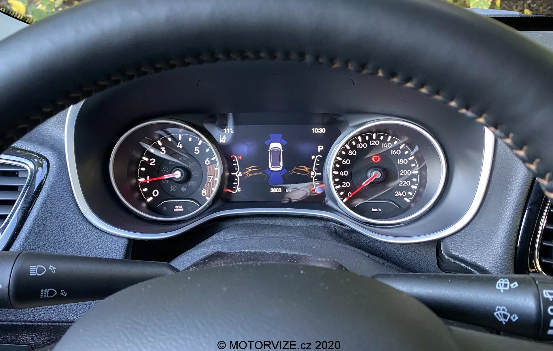 Vista aproximada do painel de instrumentos do Jeep Compass 2017 da perspectiva do motorista, exibindo indicadores analógicos e um display digital central. As características incluem um tacômetro, velocímetro de dupla unidade, indicador de seleção de marcha, medidor de combustível, hodômetro e indicadores adicionais de tecnologia de segurança, como assistência de manutenção de faixa. As costuras do volante em material semelhante ao couro destacam materiais de qualidade e conforto, enquanto as alavancas de controle e botões sugerem recursos avançados de conveniência e assistência ao motorista.