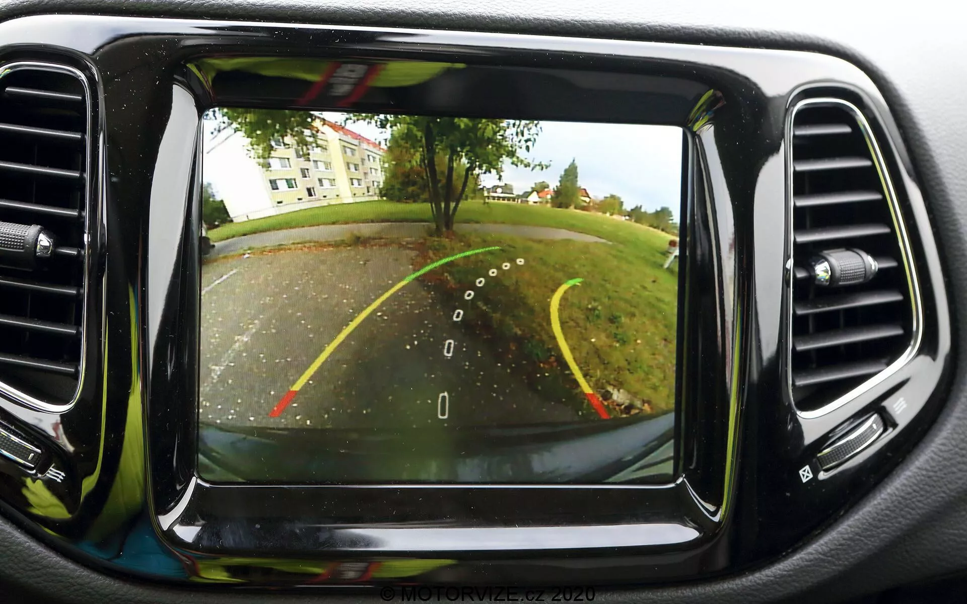 Imagem de um sistema de câmera de ré na tela do painel de um Jeep Compass 2017. A câmera, posicionada na parte traseira perto da placa, oferece uma visão em ângulo amplo atrás do veículo. São visíveis linhas de guia sobrepostas para a ré, indicando a largura do carro e a trajetória projetada com linhas amarelas e vermelhas para a distância. A cena mostra um estacionamento ou caminho de entrada com árvores e gramado ao fundo. A tela está centralmente posicionada no painel, cercada por saídas de ar condicionado, e apresenta uma qualidade de imagem clara, típica das tecnologias automotivas modernas.