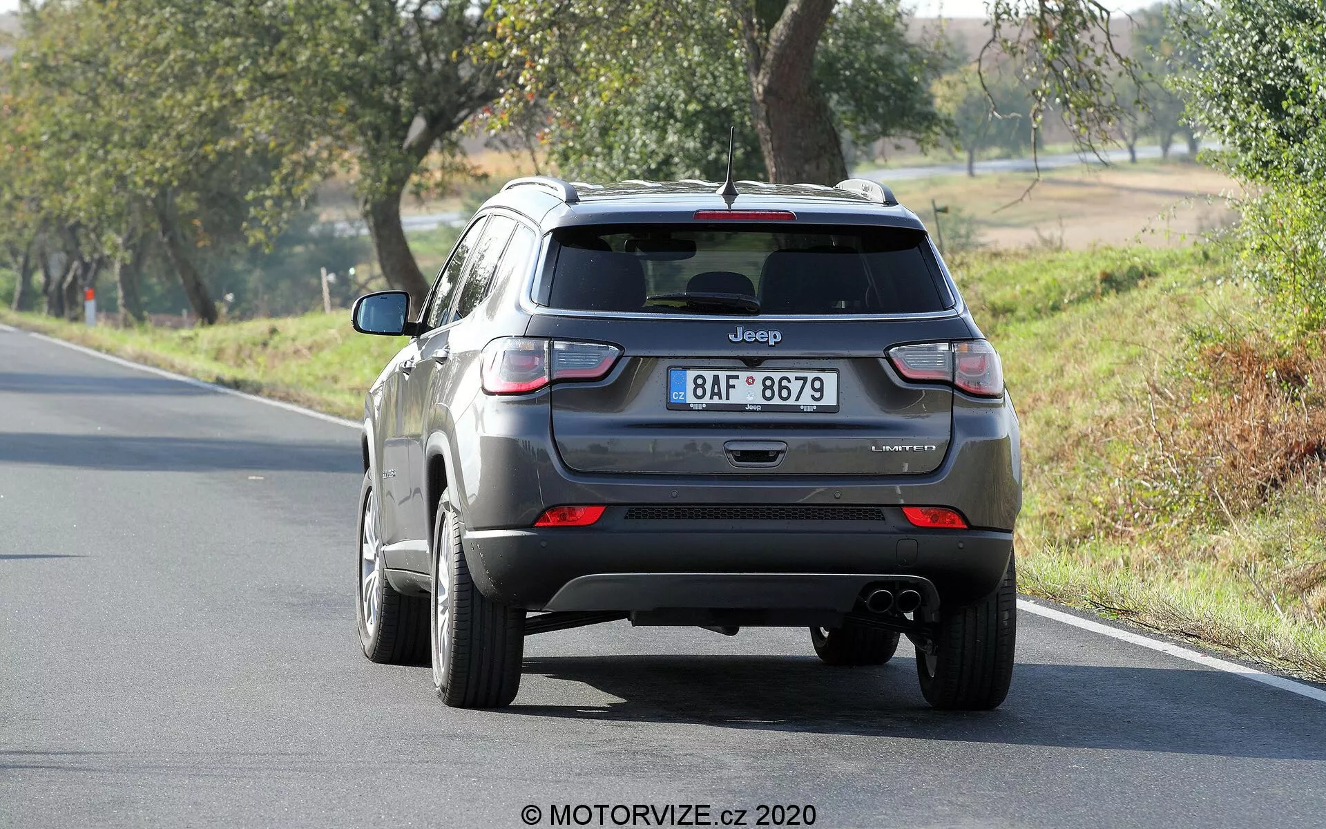 Vista traseira de um Jeep Compass prateado 2017, mostrando o porta-malas, o para-choque traseiro e as luzes traseiras. A marca 'Jeep' e o nome do modelo 'Compass' são visíveis, com o emblema do nível de acabamento ligeiramente ilegível. A imagem mostra claramente as luzes traseiras, o escapamento abaixo do para-choque e o carro posicionado em uma estrada de duas pistas cercada por árvores em um ambiente rural.