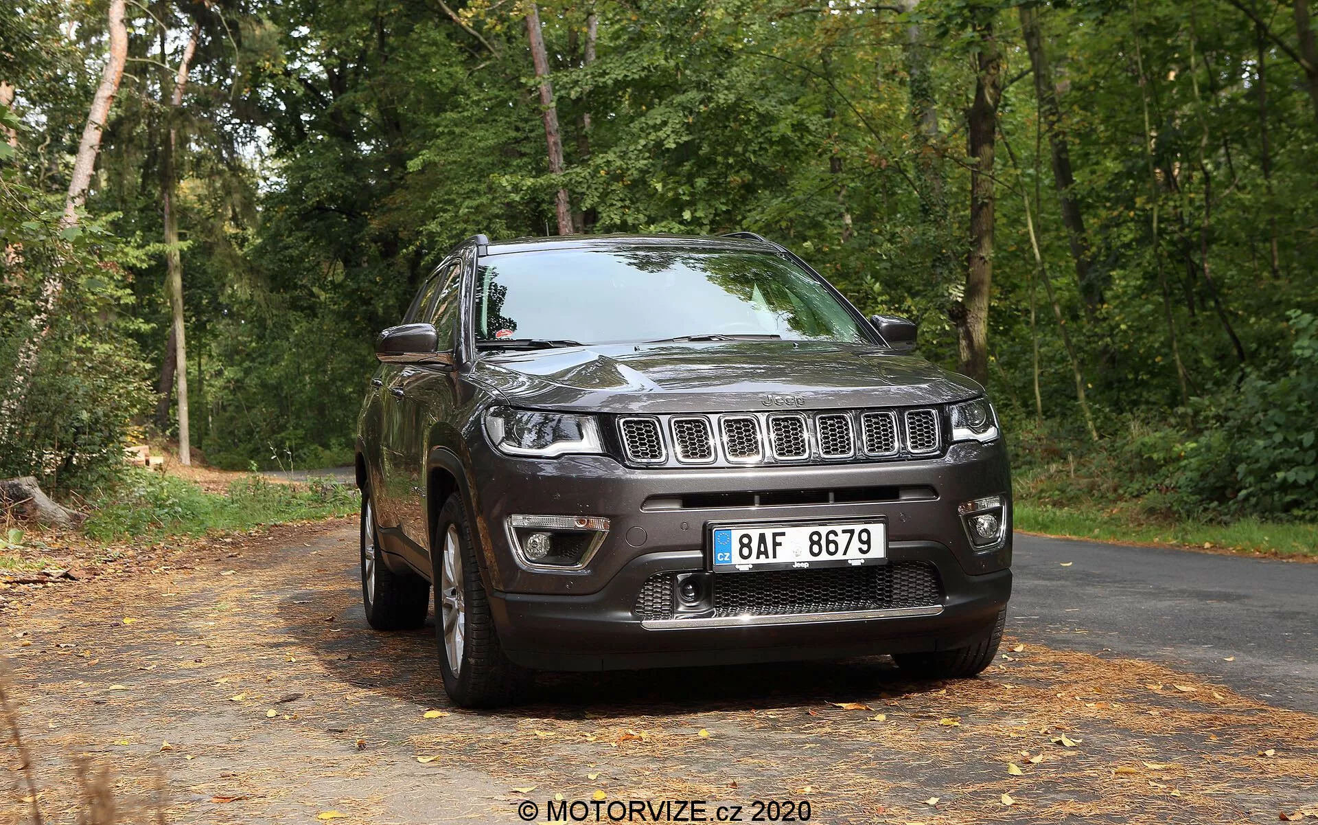Vista de três quartos da frente de um SUV Jeep Compass 2017 de cor escura em uma estrada pavimentada coberta de folhas, com árvores ao fundo em um cenário de floresta. A imagem mostra a grade frontal com ranhuras verticais distintas, ladeada por faróis que se estendem nos para-lamas, um para-choque dianteiro bem definido com faróis de nevoeiro e um capô elevado e esculpido. O perfil lateral revela rodas de liga, um para-lama visível, espelhos laterais, maçanetas das portas, contornos das janelas e um sistema de trilhos de teto, tudo iluminado por luz natural.