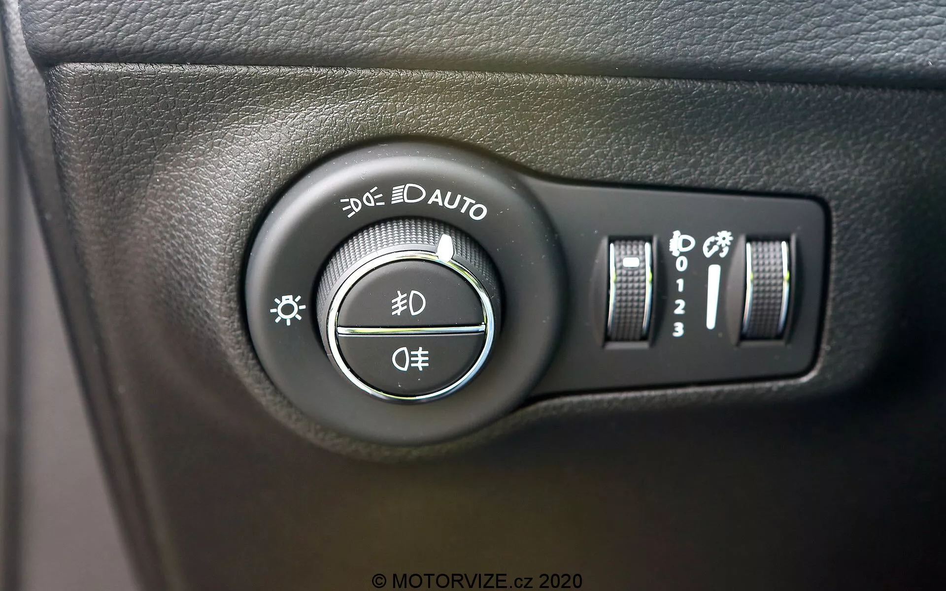 Visão detalhada do interruptor de controle do farol do Jeep Compass 2017, localizado no painel ao lado esquerdo do volante. A imagem, tirada em um ângulo ligeiro, destaca o botão rotativo com configurações para luzes de estacionamento, luzes diurnas, faróis automáticos, luzes baixas e altas, e uma posição de desligamento. Controles adicionais à direita incluem um controle deslizante vertical para o brilho do painel e um botão que pode ser para faróis altos automáticos ou sistema de nivelamento dos faróis. O interruptor está montado em uma superfície de plástico preto texturizado, com marcas brancas claras e reflexos ligeiramente brilhantes.