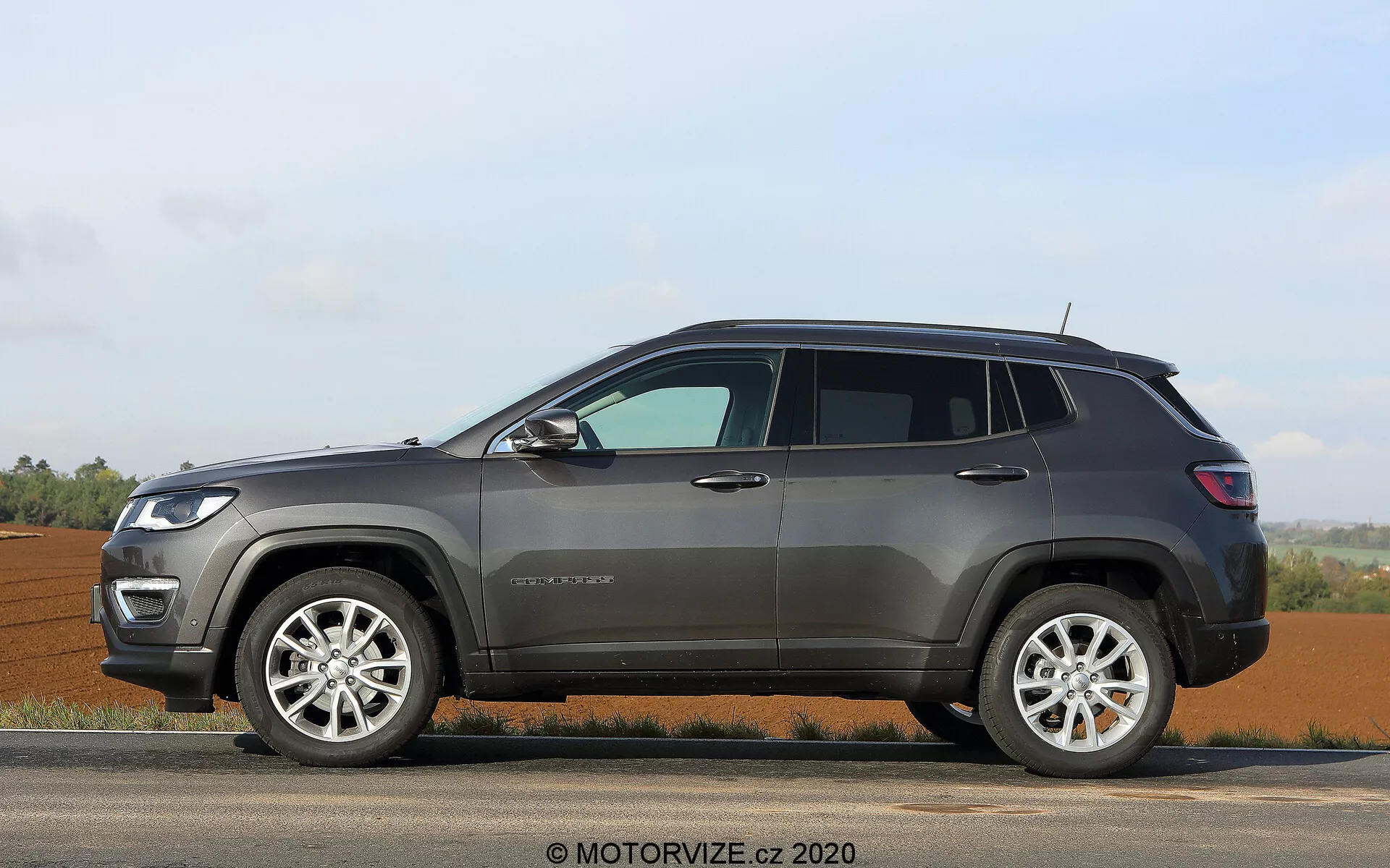 Visão de perfil lateral de um moderno SUV Jeep Compass 2017 estacionado em uma superfície nivelada, mostrando seu lado do passageiro com janelas levantadas, quatro portas, pilar B visível e vidraça traseira no pilar C. As rodas de liga leve com desenho multirraios e pneus com paredes laterais significativas são evidentes, indicando capacidade para uso em estrada e fora dela. O exterior apresenta revestimento plástico ao redor dos arcos das rodas e da parte inferior da carroceria, trilhos de teto para carga adicional, espelhos laterais provavelmente com sinalizadores integrados e contornos robustos das portas. O para-lama dianteiro exibe um emblema indicando o modelo ou nível de acabamento, e a posição elevada do veículo sugere uma alta distância ao solo, adequada para diversos terrenos. Estacionário, contra um fundo rural com campos abertos e um horizonte claro, sob condições de luz suaves.