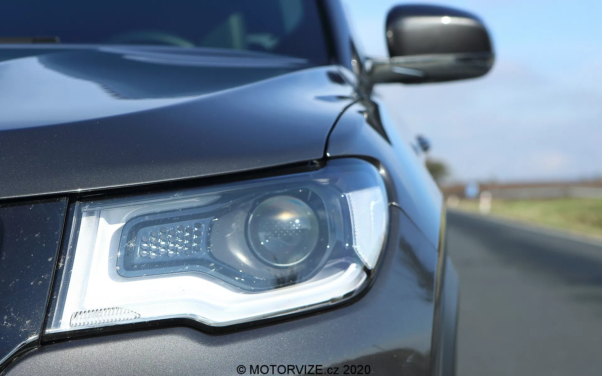 Vista de perto do lado dianteiro esquerdo de um Jeep Compass 2017, destacando o conjunto do farol a partir de um ângulo baixo ao nível do solo. O farol moderno possui um feixe projetor e luzes diurnas de LED dispostas em um padrão distinto. A carcaça do farol, elegante, integra-se perfeitamente com a carroceria de cor escura, refletindo um céu azul claro. O espelho lateral, parcialmente visível, parece combinar com a cor da carroceria e pode incluir um indicador lateral. A profundidade de campo rasa destaca os detalhes do farol e da grade frontal contra um fundo desfocado da estrada.