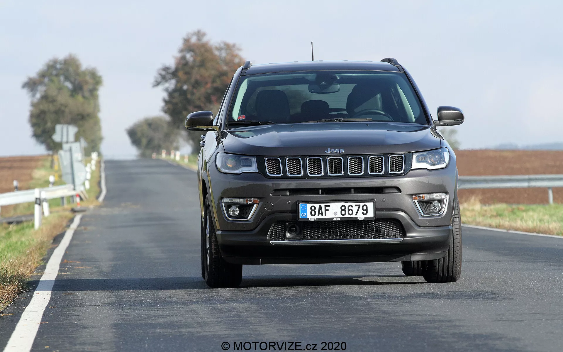 Vista frontal do Jeep Compass 2017 em movimento em uma estrada de duas faixas, capturada de um ângulo baixo, destacando a grade proeminente de sete ranhuras, os faróis quadrados com luzes diurnas modernas e as luzes de neblina e sensores de estacionamento frontais visíveis no para-choque. A estrada se estende até o horizonte com uma linha dupla amarela sólida, indicando uma zona de proibição de ultrapassagem. O fundo desfocado pelo movimento indica que o veículo está em movimento, mostrando a postura elevada típica dos SUVs.