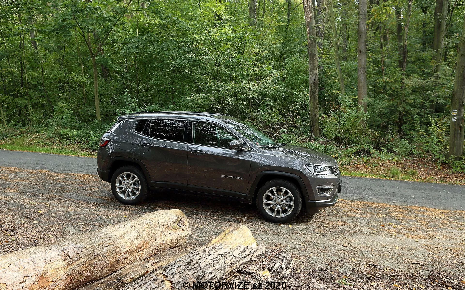Uma vista lateral de um SUV Jeep Compass 2017 estacionado em uma estrada asfaltada em um cenário natural com árvores verdes exuberantes. O veículo exibe linhas de design modernas ao longo das portas e arcos das rodas, com rodas de liga de múltiplos raios visíveis. Ele tem uma alta altura do solo, sugerindo capacidades off-road. A imagem captura parte da grade frontal e dos faróis, indicando um estilo contemporâneo. Barras de teto, sinais de viragem integrados nos espelhos laterais e janelas escurecidas estão presentes. A pintura de cor escura com um acabamento brilhante indica um veículo bem conservado. Enquanto estacionado, o SUV é mostrado com abundante luz natural, ideal para apresentar suas características e apelo para entusiastas de atividades ao ar livre e aventuras.