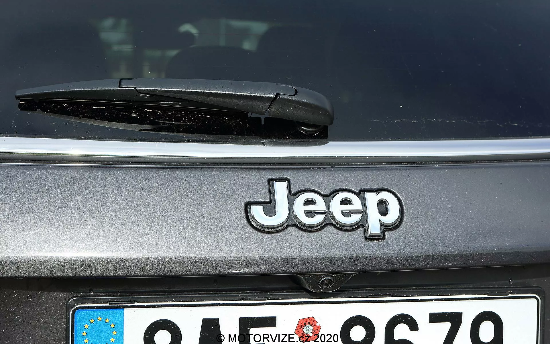Imagem em close-up da parte traseira de um Jeep Compass 2017, mostrando o logotipo centralizado da Jeep, o limpador do para-brisas traseiro e a placa de estilo europeu parcialmente visível com o identificador do país desfocado