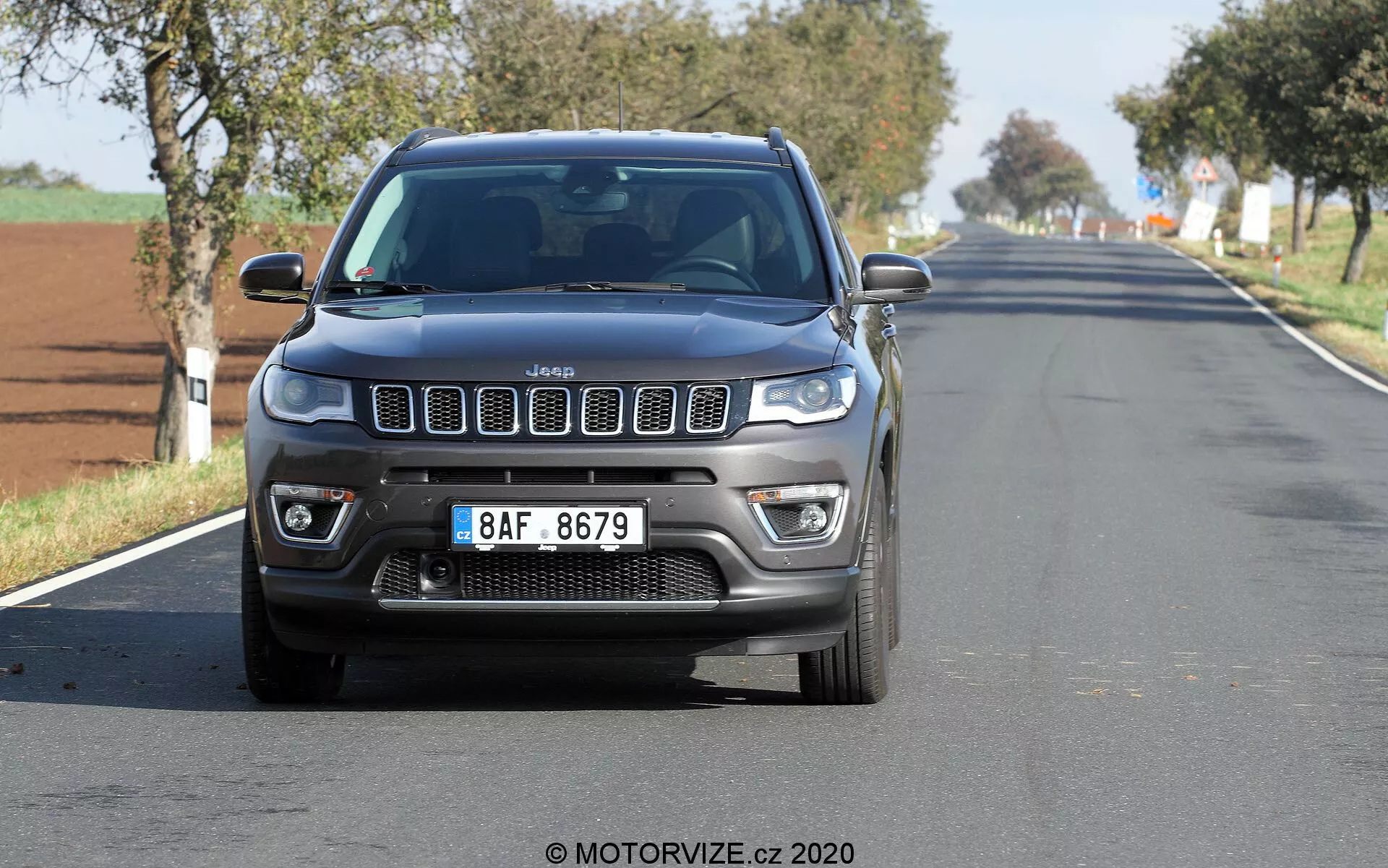 Vista frontal de um Jeep Compass 2017 viajando em uma estrada rural ou suburbana de duas pistas, capturada de um ângulo baixo, mostrando sua icônica grade de sete fendas, DRLs de LED, faróis de neblina e contornos robustos do capô. O carro está centralizado e relativamente próximo à câmera, com um leve desfoque nas rodas e no fundo. Céu limpo sugere um dia claro e ensolarado.
