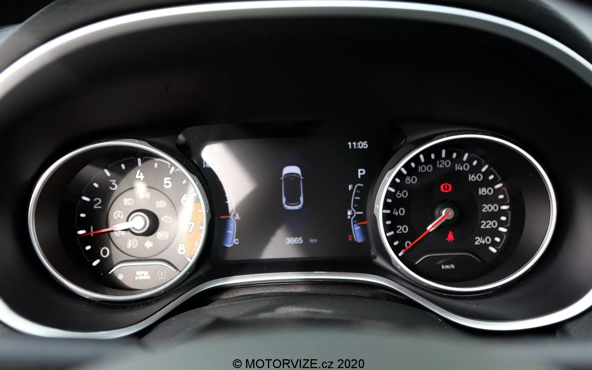 Vista de perto do painel de instrumentos do Jeep Compass 2017 com uma combinação de mostradores analógicos e um display digital. À esquerda, um velocímetro analógico marcado até 260 com um ponteiro apontando logo acima de 0. O display digital central inclui uma representação gráfica do carro com sensores e duas portas abertas, indicando assistência de estacionamento e status do veículo. Um relógio mostra 11:05. À direita, há um tacômetro analógico com zona vermelha começando antes de 7, marcado até 8 (incrementos de 1.000 rpm). Luzes de aviso adicionais e ícones são visíveis, tornando esta uma representação bem iluminada e clara das leituras modernas e tradicionais do veículo e das tecnologias de assistência ao condutor.