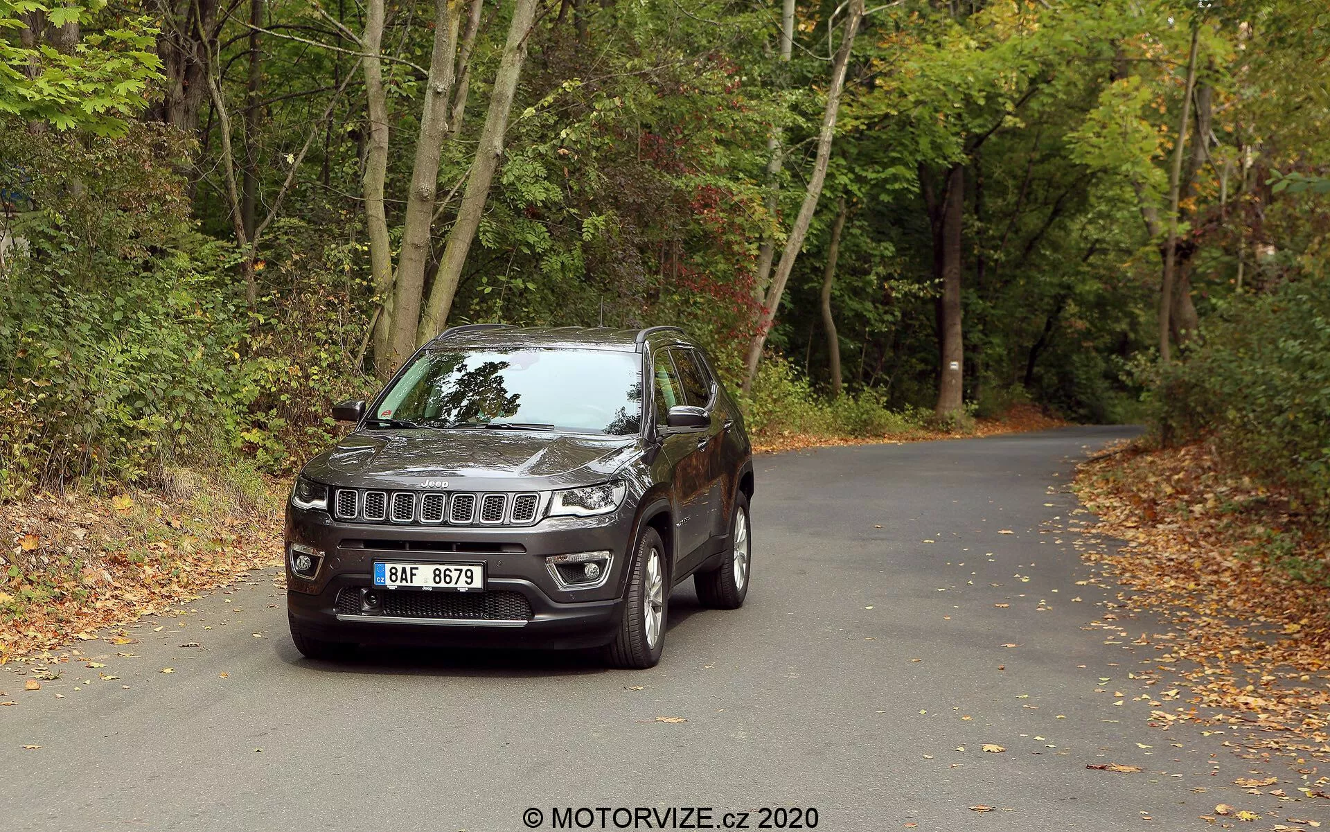 Um SUV Jeep Compass 2017 posicionado em uma estrada de asfalto rodeada por florestas com folhas no chão, sugerindo que é outono. O veículo é fotografado de um ângulo de três quartos frontais, exibindo a distintiva grade de sete fendas da Jeep e o emblema central, faróis modernos de LED e luzes de nevoeiro integradas no para-choque. Possui rodas de liga, espelhos laterais com sinais de seta integrados, uma construção robusta com revestimento dos arcos das rodas e vidros ligeiramente escurecidos. A vista detalhada destaca elementos de design e tecnológicos, sugerindo sua adequação para aventuras urbanas e off-road.