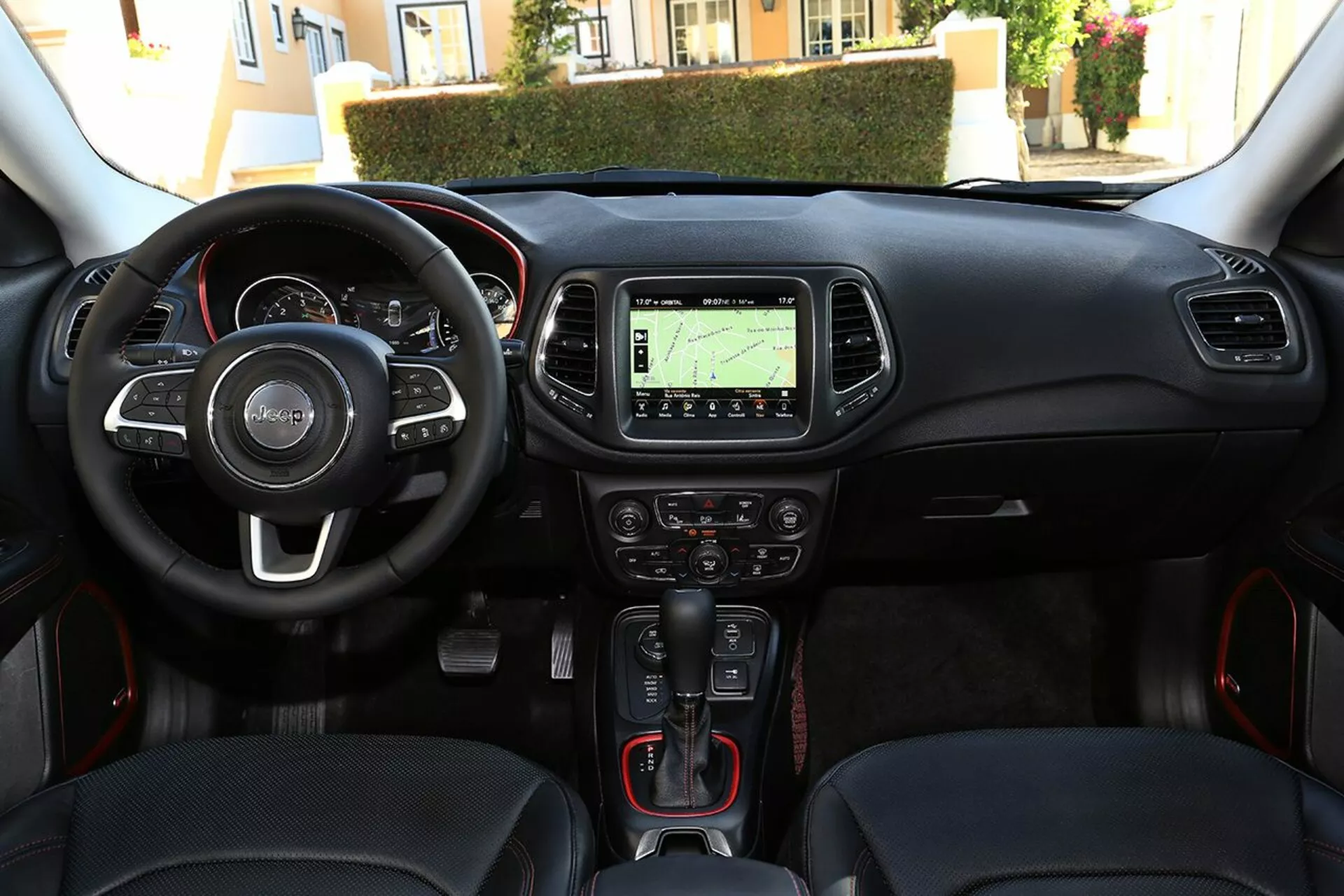 Vista interior do Jeep Compass 2017 a partir da posição central do banco traseiro, destacando o volante moderno com controles multifuncionais, cluster de instrumentos avançado, tela touchscreen central com navegação, transmissão automática com modo manual, design elegante do painel com iluminação ambiente, bancos dianteiros contornados com costura vermelha, e controles elétricos de janela na porta do motorista.