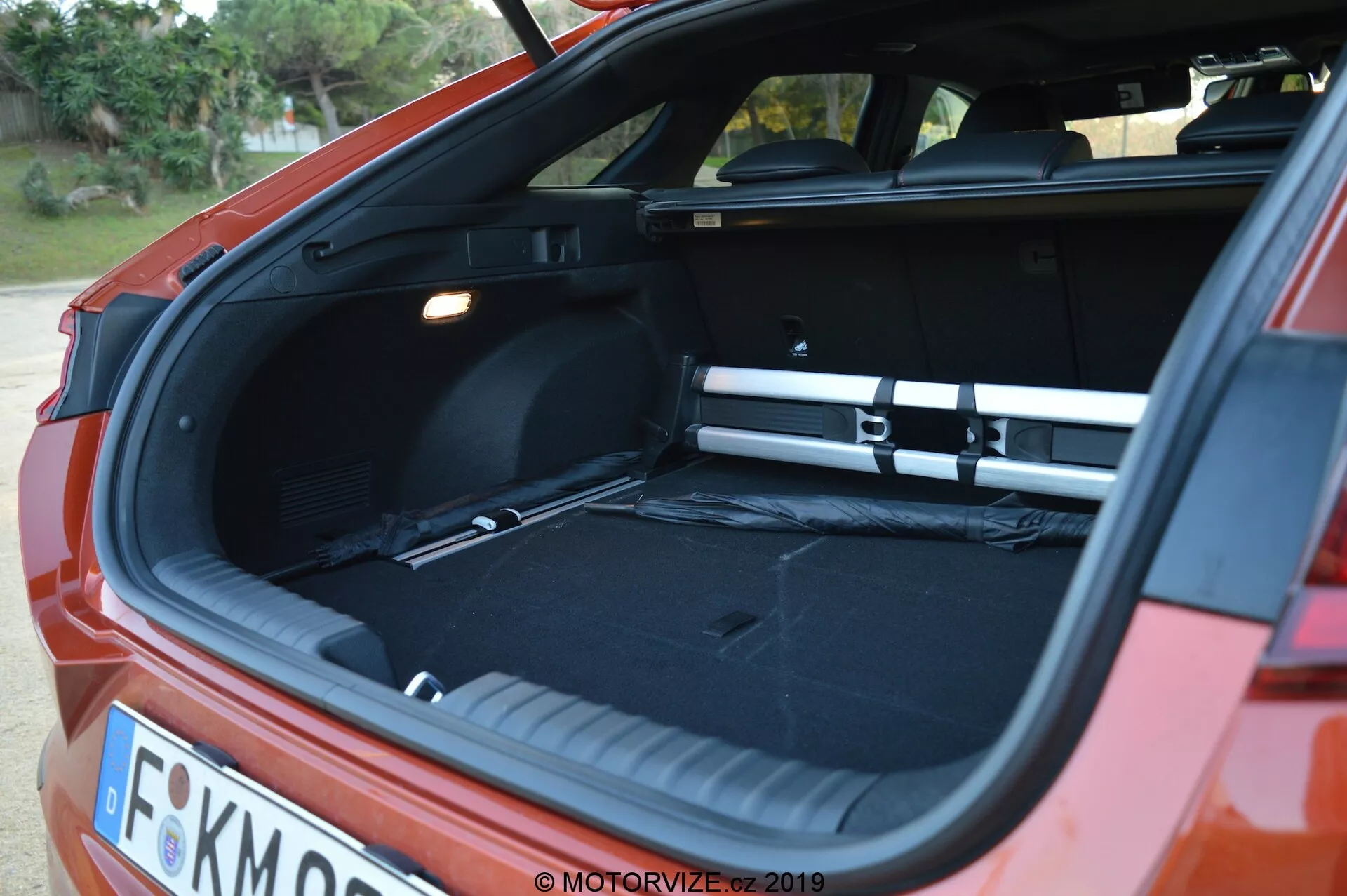 A imagem captura uma vista traseira angulada do Kia Ceed 2021 vermelho, focando na tampa traseira hatchback aberta. É amanhecer ou anoitecer, e a iluminação suave destaca o espaçoso porta-malas vazio com interior preto. Uma cobertura de carga está no lugar, estendendo-se por toda a largura do porta-malas. A foto, tirada do canto traseiro com um leve ângulo lateral, também mostra partes do para-choque traseiro, luzes traseiras e uma placa de matrícula parcialmente visível. O porta-malas tem um piso plano de carga, uma fonte de luz à esquerda e um compartimento lateral à direita.