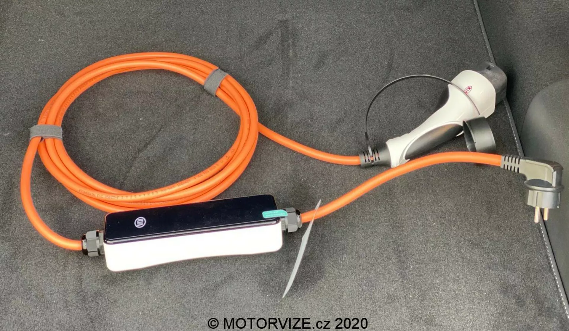 Cabo de carregamento de veículo elétrico (EV) e configuração de adaptador para o Kia Ceed 2018, com um EVSE (Equipamento de Abastecimento de Veículo Elétrico) portátil laranja de alta visibilidade para segurança. A configuração inclui um conector e plugue de carregamento para uso em tomada doméstica, bem como uma caixa de controle em linha com indicadores LED, tudo disposto no chão acarpetado do porta-malas ou área de carga do veículo.