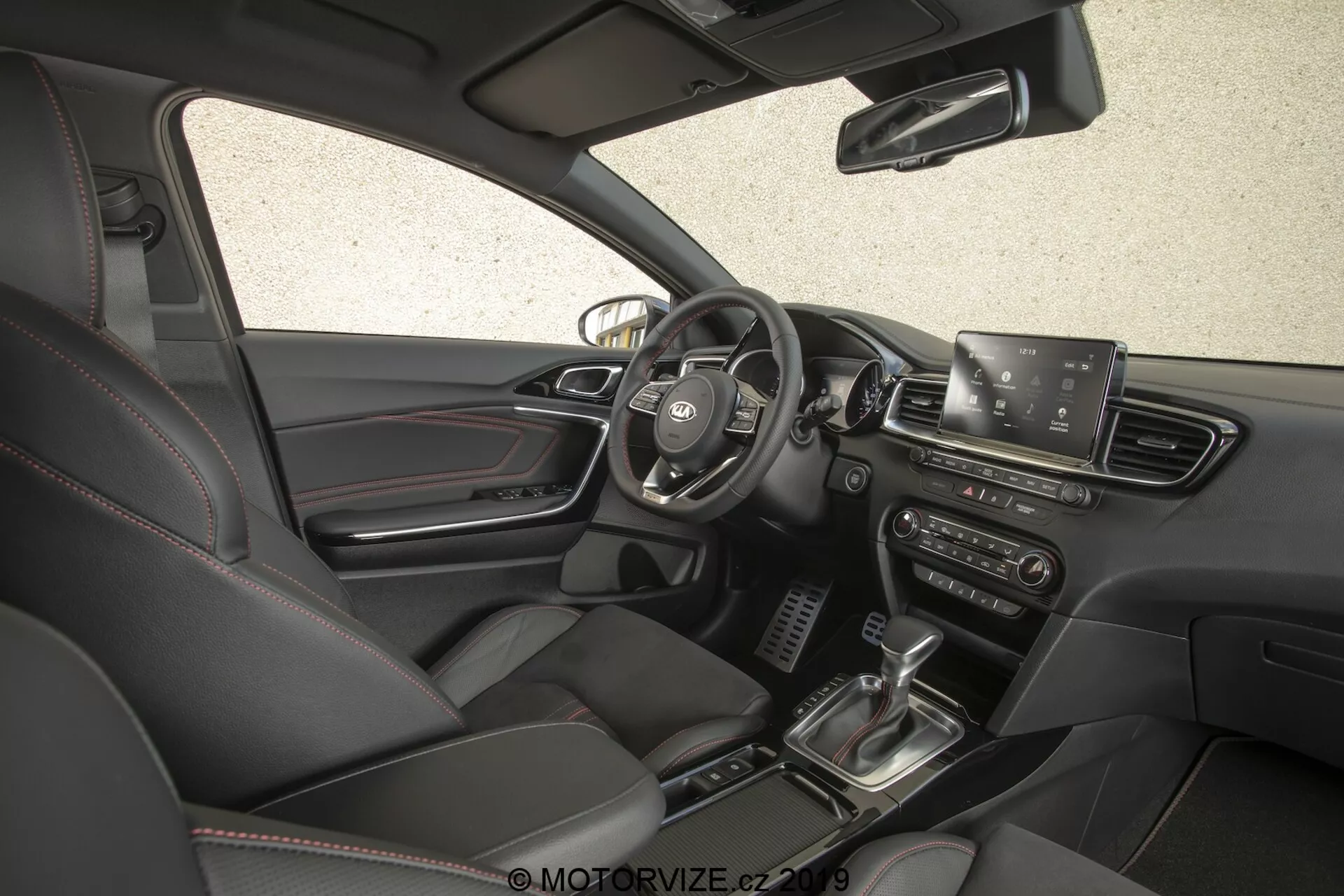 Vista interior do Kia Ceed 2021, mostrando o lado do condutor a partir de um ângulo alto e ligeiramente voltado para a frente. A imagem, capturada do lado do passageiro, oferece uma visão completa do painel, incluindo o volante central com botões de controle, visibilidade parcial do painel de instrumentos, uma tela de infotainment proeminente, saídas de ar, botões e dials de controle do clima, alavanca de câmbio, sistema de partida por botão, assentos de couro com controle de ajuste elétrico e uma paleta de cores interiores escuras.