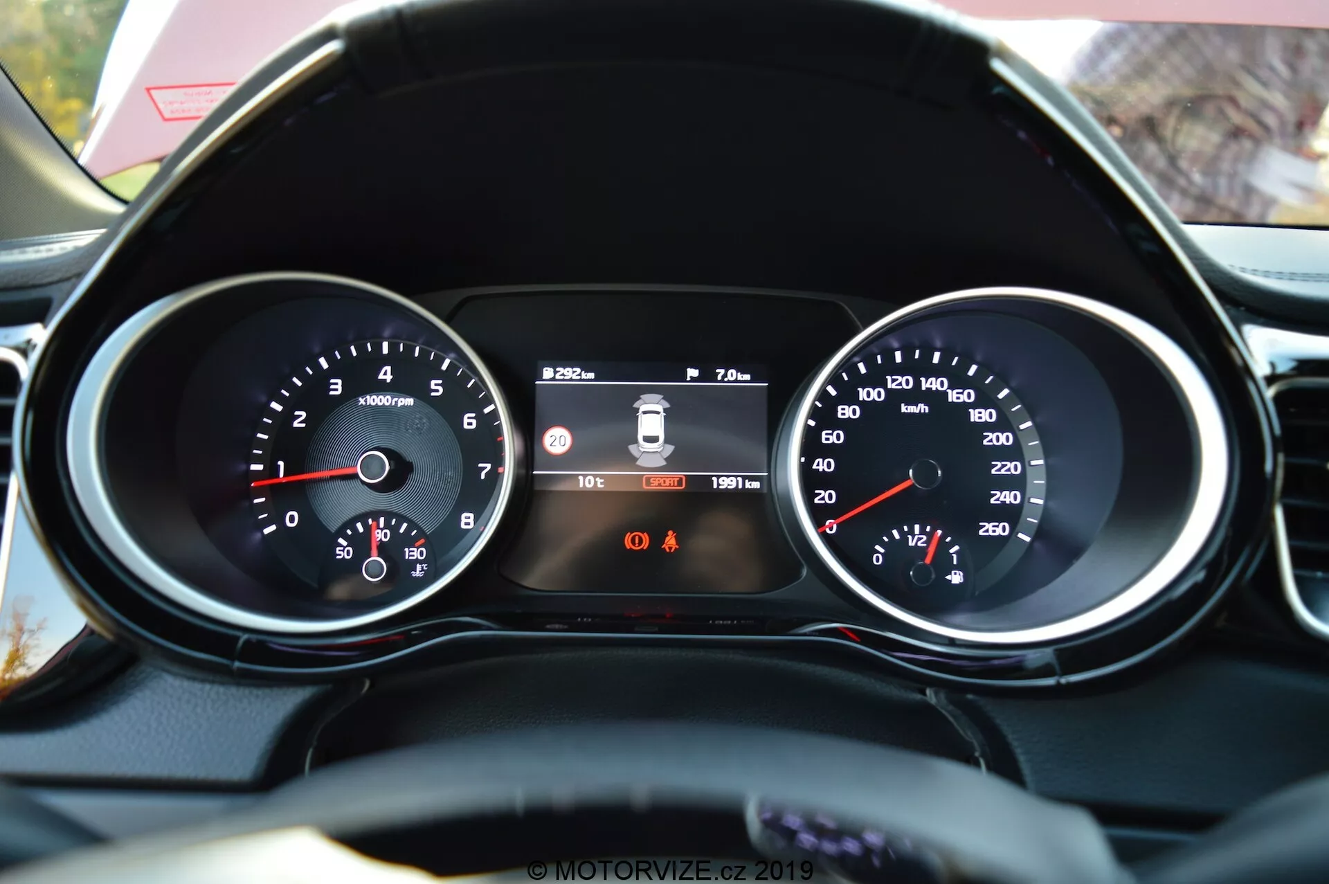 Close-up da perspectiva do condutor do painel de instrumentos do Kia Ceed 2021 e parte do volante com acabamento em couro. As características incluem um tacômetro à esquerda com leituras de RPM, um display digital central mostrando o sistema de aviso de saída de faixa, a hora e o odômetro, e um velocímetro à direita com a velocidade em km/h. Abaixo do display central, duas luzes indicadoras de aviso são visíveis, sendo identificável o lembrete do cinto de segurança.
