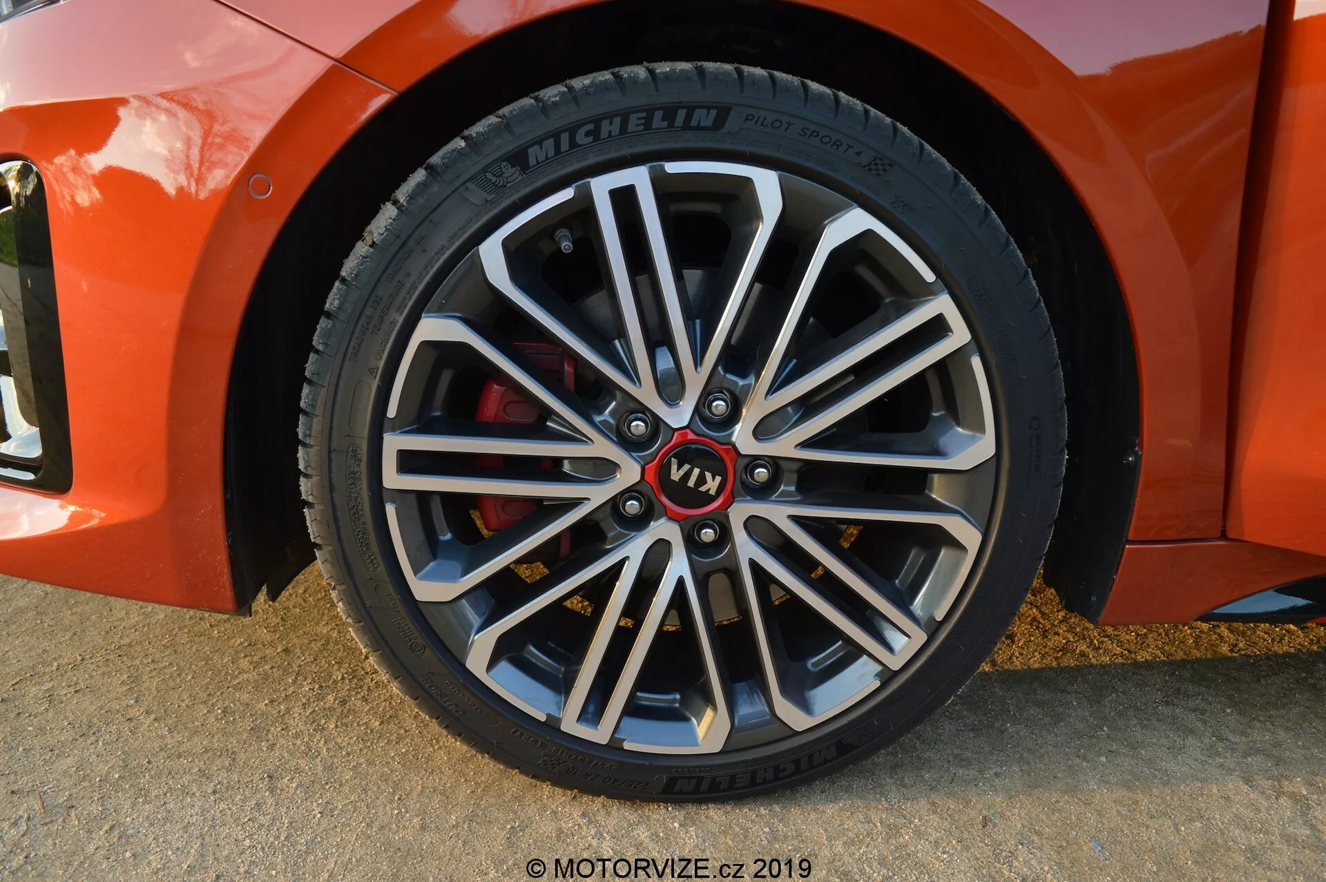 Close-up da roda dianteira do lado do passageiro do Kia Ceed 2021, em ângulo levemente diagonal, mostrando uma roda de liga leve de vários raios com seções internas mais escuras e bordas externas brilhantes. Apresenta cubo da roda com marcação vermelha 'VT1', pneu Michelin com informações detalhadas na parede lateral e pinça de freio vermelha indicando componentes orientados para desempenho. As sombras suaves e os destaques sugerem que a foto foi tirada em luz natural, provavelmente durante a hora dourada.