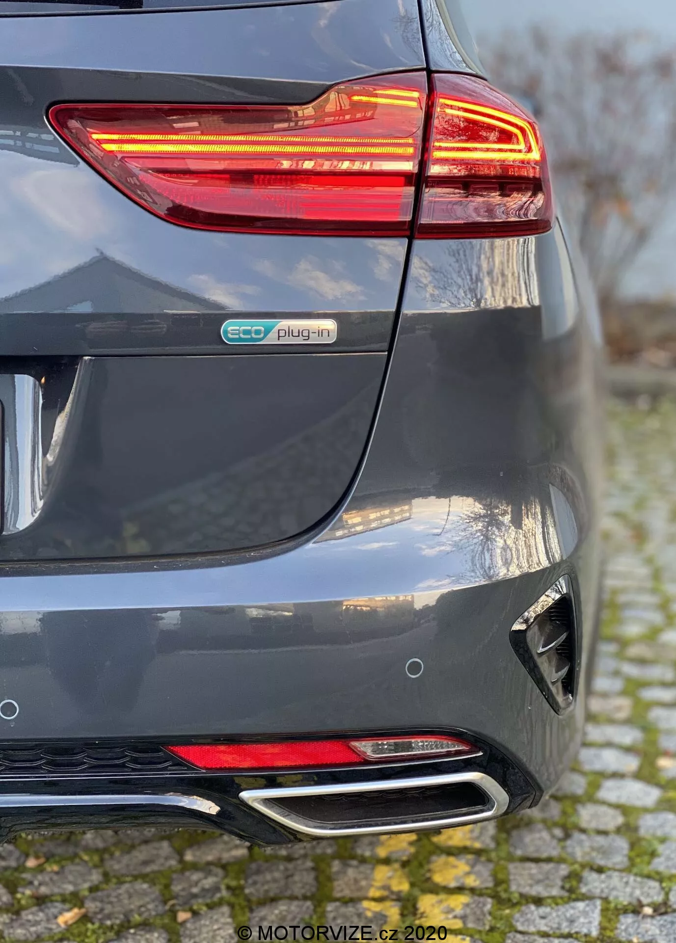 Vista detalhada da seção traseira de um Kia Ceed 2018 em cinza metálico, destacando a luz traseira LED iluminada, o emblema eco plug-in, o para-choque traseiro esculpido com refletor e difusor, e as pontas duplas cromadas do escapamento. A perspectiva mostra a convergência do para-choque traseiro e do lado direito, enfatizando os elementos do veículo híbrido elétrico e as características de design moderno.