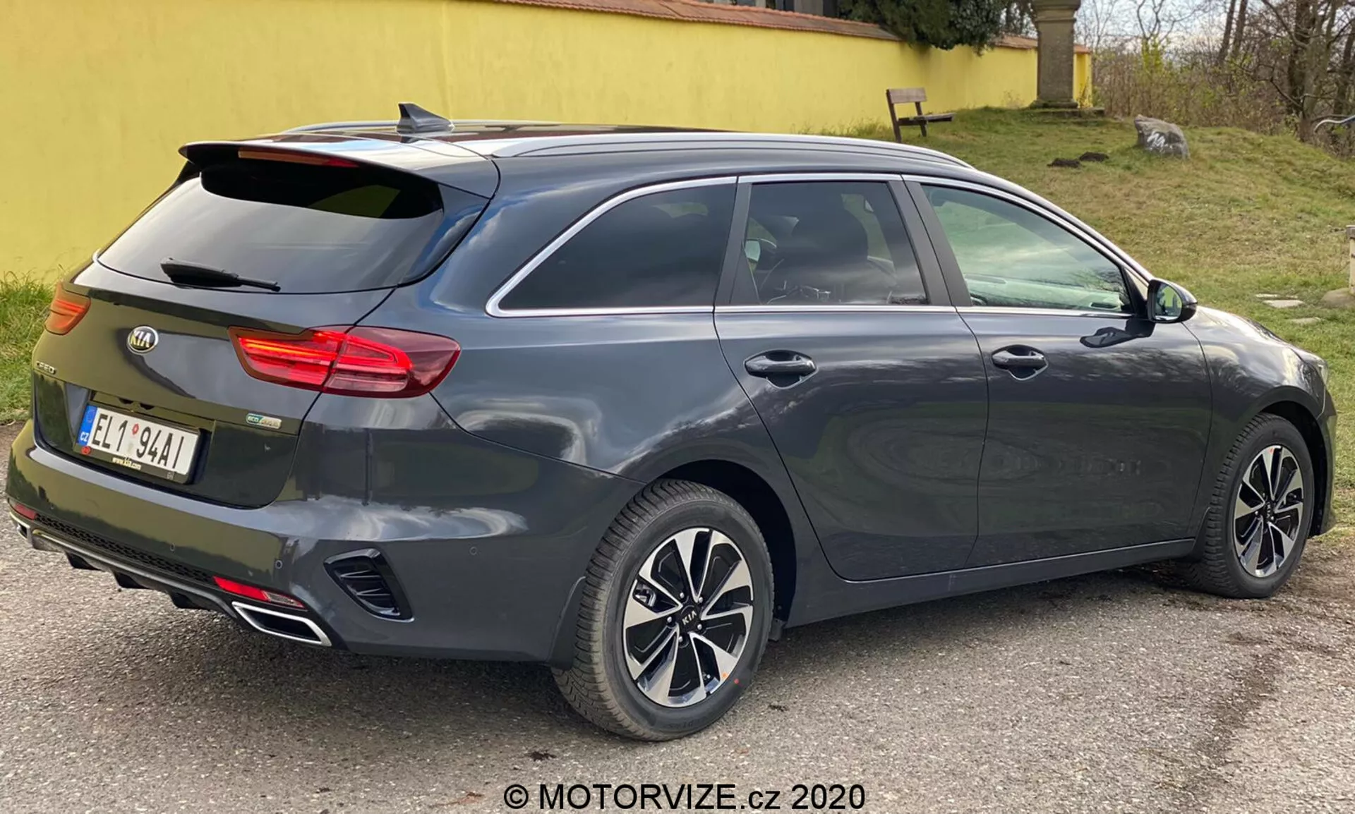 Um Kia Ceed 2018 metálico cinza escuro, mostrado a partir de uma vista traseira de três quartos, destacando seu design moderno de hatchback ou perua com janela traseira inclinada, spoiler traseiro integrado, rodas de liga leve de vários raios, maçanetas das portas e espelhos laterais na cor da carroceria provavelmente com sinais de seta integrados, conjunto de luzes traseiras horizontais distintivo possivelmente com tecnologia LED, antena tipo barbatana de tubarão, ponteiras duplas de escape, terceira luz de freio montada em altura, capacidade de descongelamento da janela traseira, sensores ou refletores indicativos de um sistema de assistência ao estacionamento traseiro, fotografado de perto com iluminação neutra.
