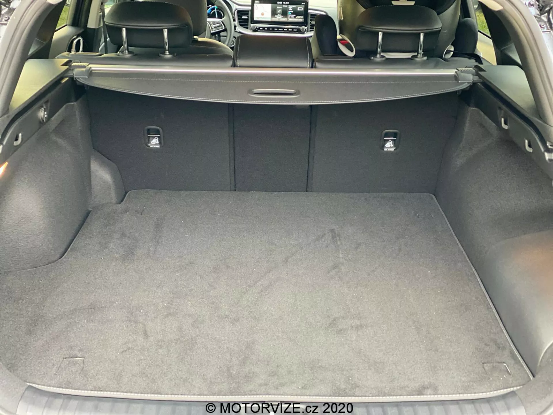 Vista detalhada do porta-malas do Kia Ceed 2018 de uma perspectiva traseira, destacando a área de carga espaçosa e limpa com uma superfície acarpetada. A porta do porta-malas está levantada, revelando uma cobertura retrátil, encostos dos bancos traseiros de cor mais escura, possivelmente de couro, e abas prateadas para dobrar os assentos. As laterais são revestidas com um material macio e há um pequeno compartimento visível no lado direito. A imagem é tirada de um ângulo leve, mostrando o design e os materiais do interior do porta-malas do carro.