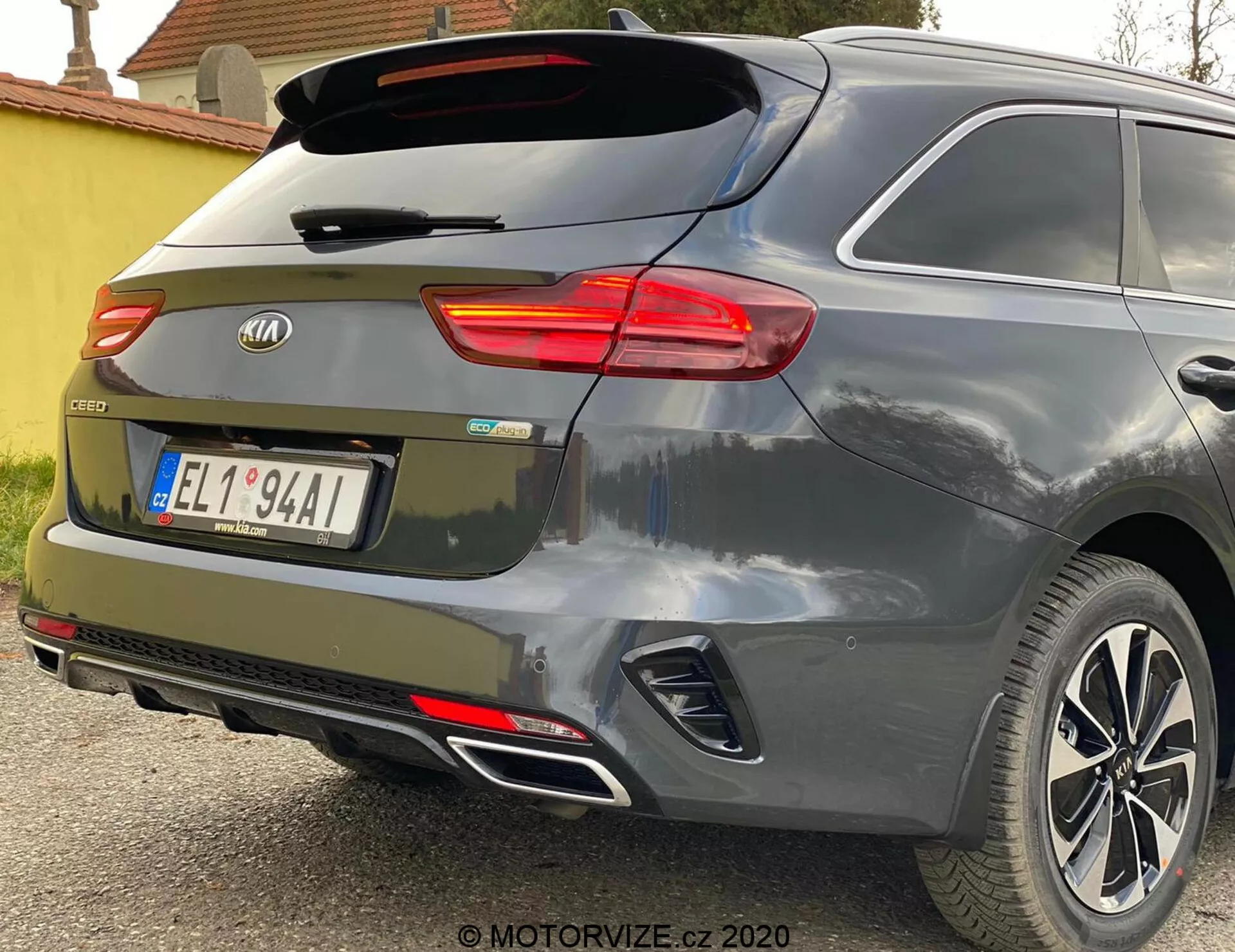 Vista parcial do ângulo traseiro de um Kia Ceed 2018, destacando a luz traseira direita iluminada, o para-brisa traseiro com limpador, o porta-malas com o emblema da marca e a insígnia do modelo 'CEED', refletores integrados no para-choques traseiro, sensores de estacionamento, sistema de escape duplo e rodas de liga leve contra uma pintura metálica cinza ou prata.