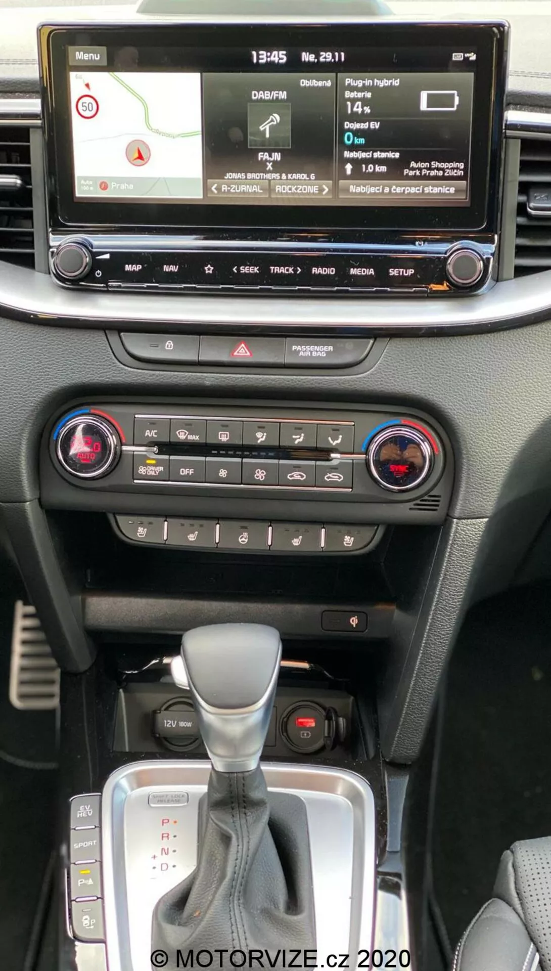 Interior do Kia Ceed 2018 mostrando o console central da perspectiva do passageiro na fila da frente, com tela de infoentretenimento digital com mapa de navegação, reprodutor de mídia, configurações do veículo, controles de clima com sistema de zona dupla, botão de luzes de emergência, alavanca de câmbio automático com proteção de couro e slot para chave de partida sem chave.