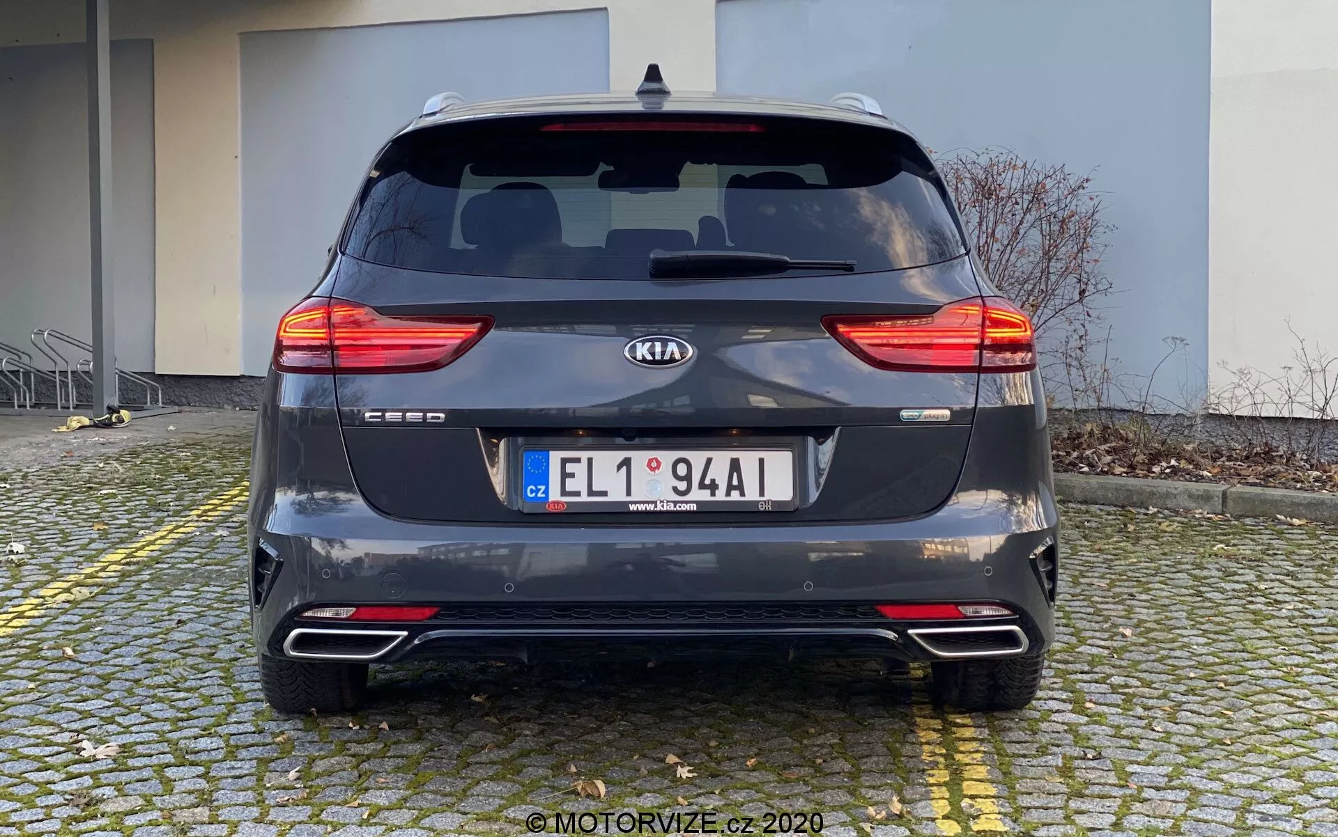 Vista traseira de um Kia Ceed 2018 estacionado em um piso de paralelepípedos perto de uma calçada. O carro, capturado durante o dia de um ângulo direto ao nível dos olhos, exibe suas lanternas traseiras de LED divididas, para-brisa traseiro com limpador, emblema do fabricante, nome do modelo, placa de licença, para-choque traseiro com refletores e sensores de estacionamento, sistema de escape duplo e acabamento metálico cinza escuro. O fundo inclui um prédio, indicando uma área residencial ou comercial.