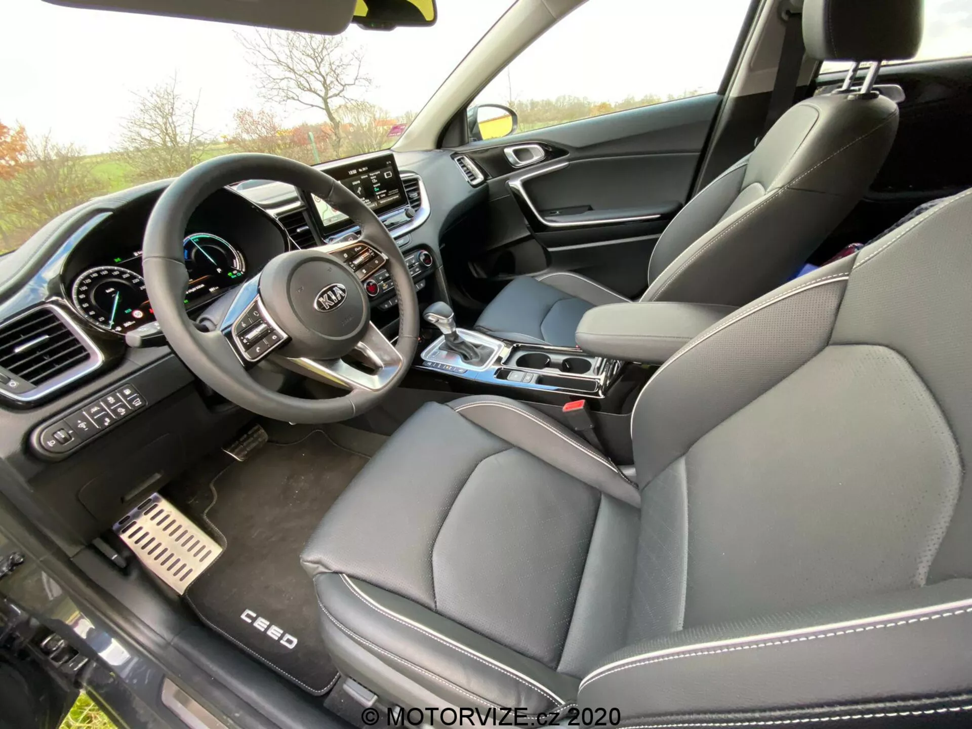 Imagem do interior de um Kia Ceed 2018 a partir da perspectiva do passageiro, capturando o compartimento frontal com foco no painel de instrumentos, console central e banco do motorista. O volante com múltiplos botões de controle e o logotipo da Kia é visível, juntamente com um painel de instrumentos digital parcialmente visível. A parte central do painel contém uma grande tela touchscreen de infotainment cercada por controles manuais de climatização. O console central inclui uma alavanca de câmbio, porta-copos e botões de controle. O banco dianteiro do passageiro possui estofamento em couro e o painel da porta é composto por materiais mistos, sugerindo um nível de acabamento luxuoso. Os pedais de freio e acelerador em metal também indicam um toque esportivo premium.