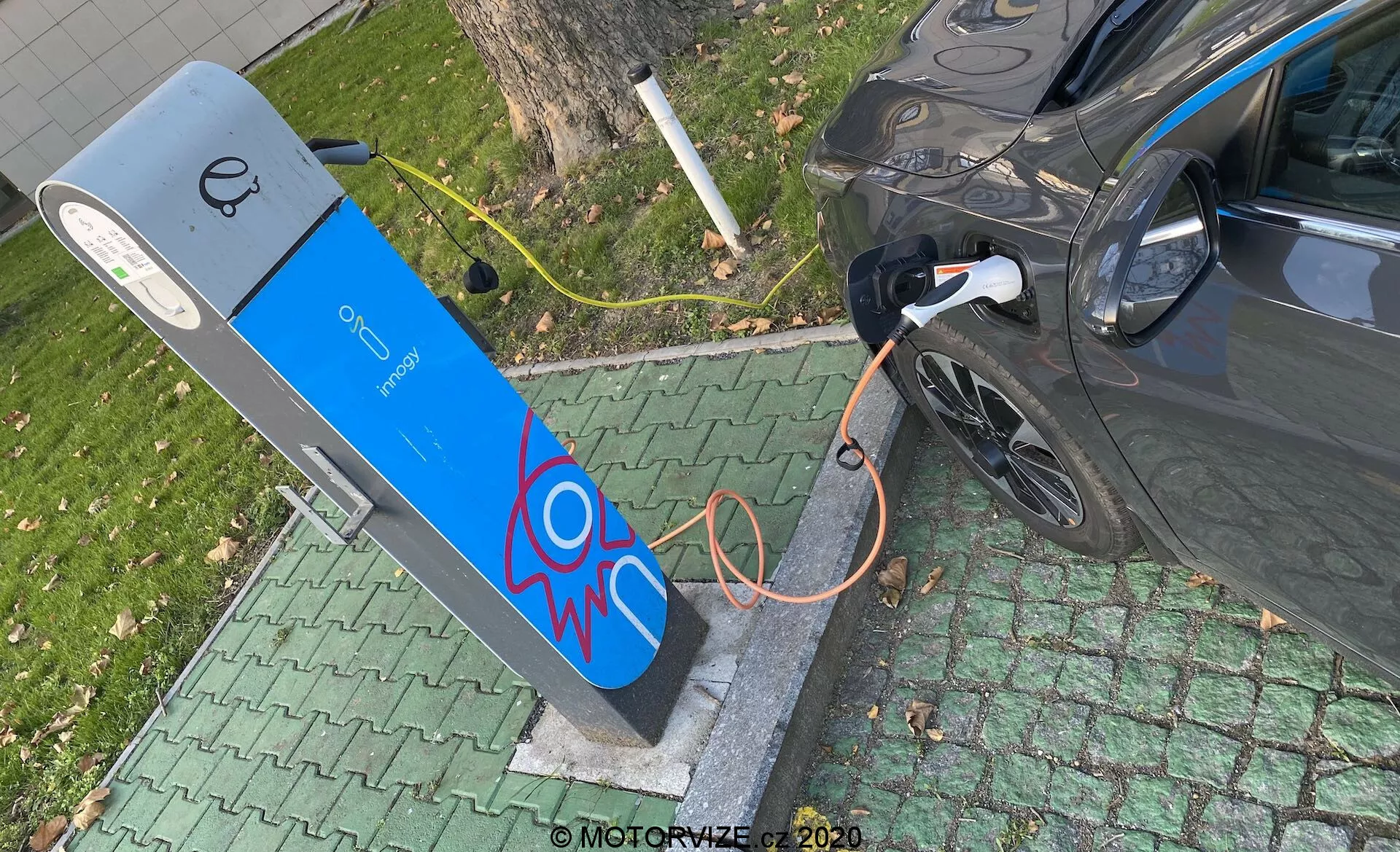 Vista aérea de um veículo elétrico moderno a ser carregado numa estação de carregamento pública. A imagem captura a seção traseira direita do carro, focando no porto de carregamento com o cabo laranja grosso conectado. A estação de carregamento apresenta uma fachada azul com um gráfico de faísca elétrica e um ecrã no topo, sugerindo elementos de interface de usuário. A cena está situada num ambiente verde semelhante a um parque, indicando uma localização urbana ou suburbana conveniente.