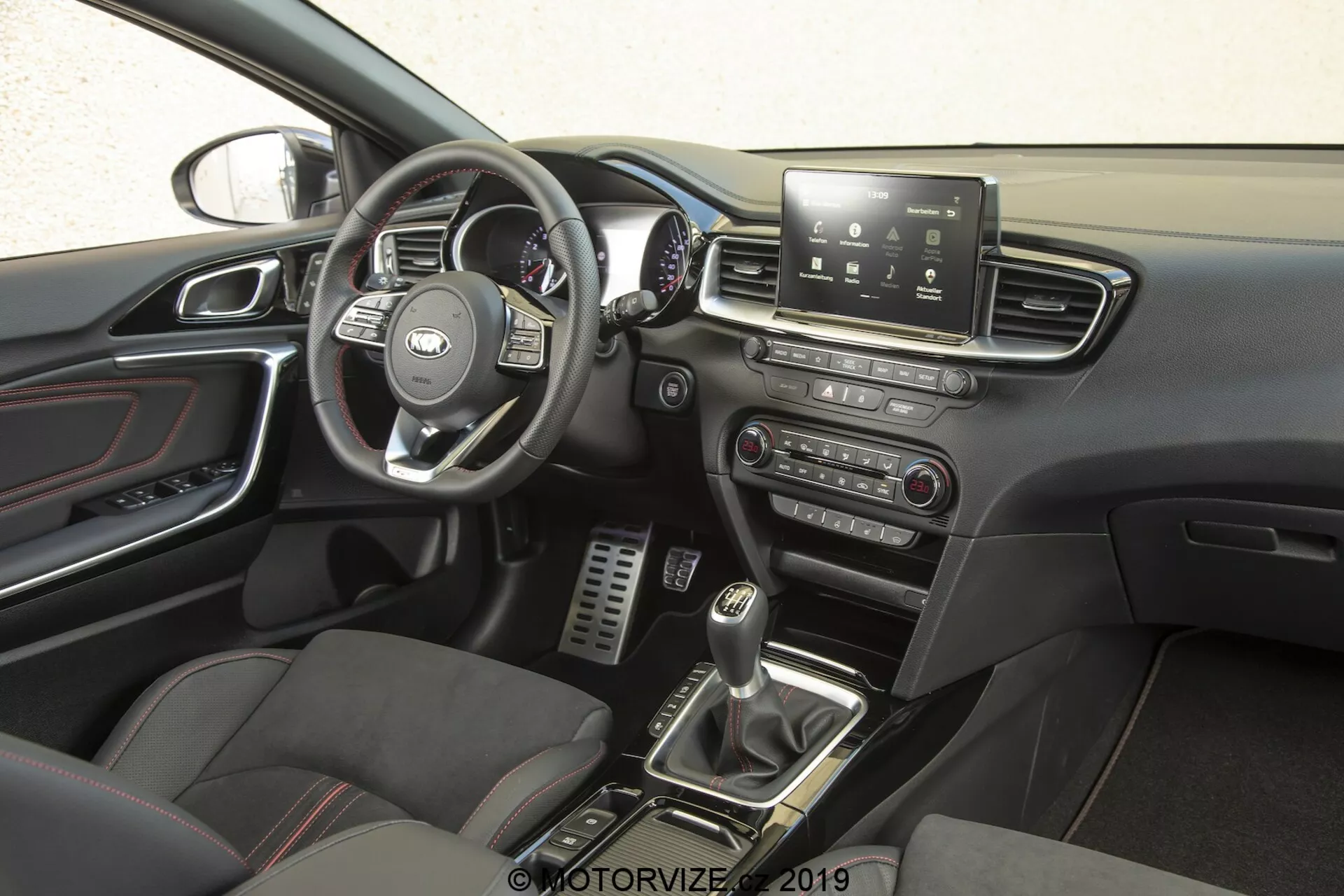 O interior do Kia Ceed 2021, focado no lado do motorista a partir de uma perspectiva lateral. O volante, com o logotipo reconhecível do fabricante, possui controles multifuncionais para gerenciar o sistema de infoentretenimento, controle de cruzeiro, conectividade telefônica e possivelmente comandos de voz. Atrás, é possível ver o velocímetro, o tacômetro e os medidores de informações do motorista. O console central possui uma tela de toque para infoentretenimento que oferece navegação, configurações do veículo, reprodução de mídia e conectividade Bluetooth. Abaixo, há controles manuais para os sistemas de áudio e climatização. A alavanca de câmbio de transmissão manual, cercada por botões para modos de condução ou controle de tração, e os assentos esportivos com inserções de couro e tecido, aumentam a sensação premium e orientada para o desempenho. Pedais metálicos no espaço para os pés do motorista sugerem uma ênfase no desempenho, enquanto os detalhes cromados nas saídas de ventilação, maçanetas das portas e outros detalhes adicionam sofisticação. O painel da porta inclui controles para os vidros elétricos e possivelmente ajustes dos espelhos elétricos, e uma parte do retrovisor é visível, sugerindo recursos como escurecimento automático ou uma bússola integrada.