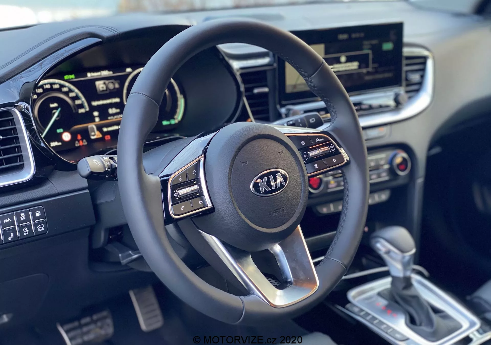 Vista em primeira pessoa do interior do Kia Ceed 2018 destacando o volante multifuncional moderno de três raios com logotipo da Kia e o painel de instrumentos digital, mostrando velocímetro e possível tacômetro, juntamente com o sistema de infoentretenimento touchscreen, controles de clima, alavanca de câmbio automática prateada e console central detalhado.