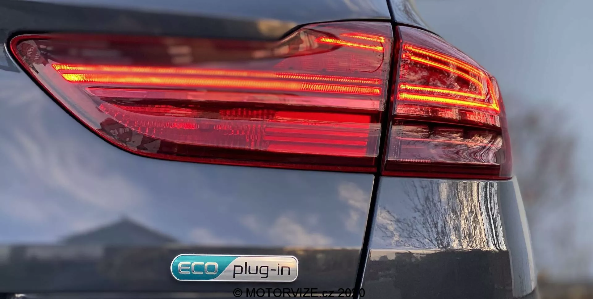 Vista aproximada do lado traseiro esquerdo de um Kia Ceed 2018, com foco no conjunto de luzes traseiras iluminadas com design multicamadas. O emblema 'ECO plug-in' indica que é um veículo híbrido plug-in. O acabamento brilhante e a tampa do porta-malas ligeiramente visível adicionam detalhes ao design geral do carro.