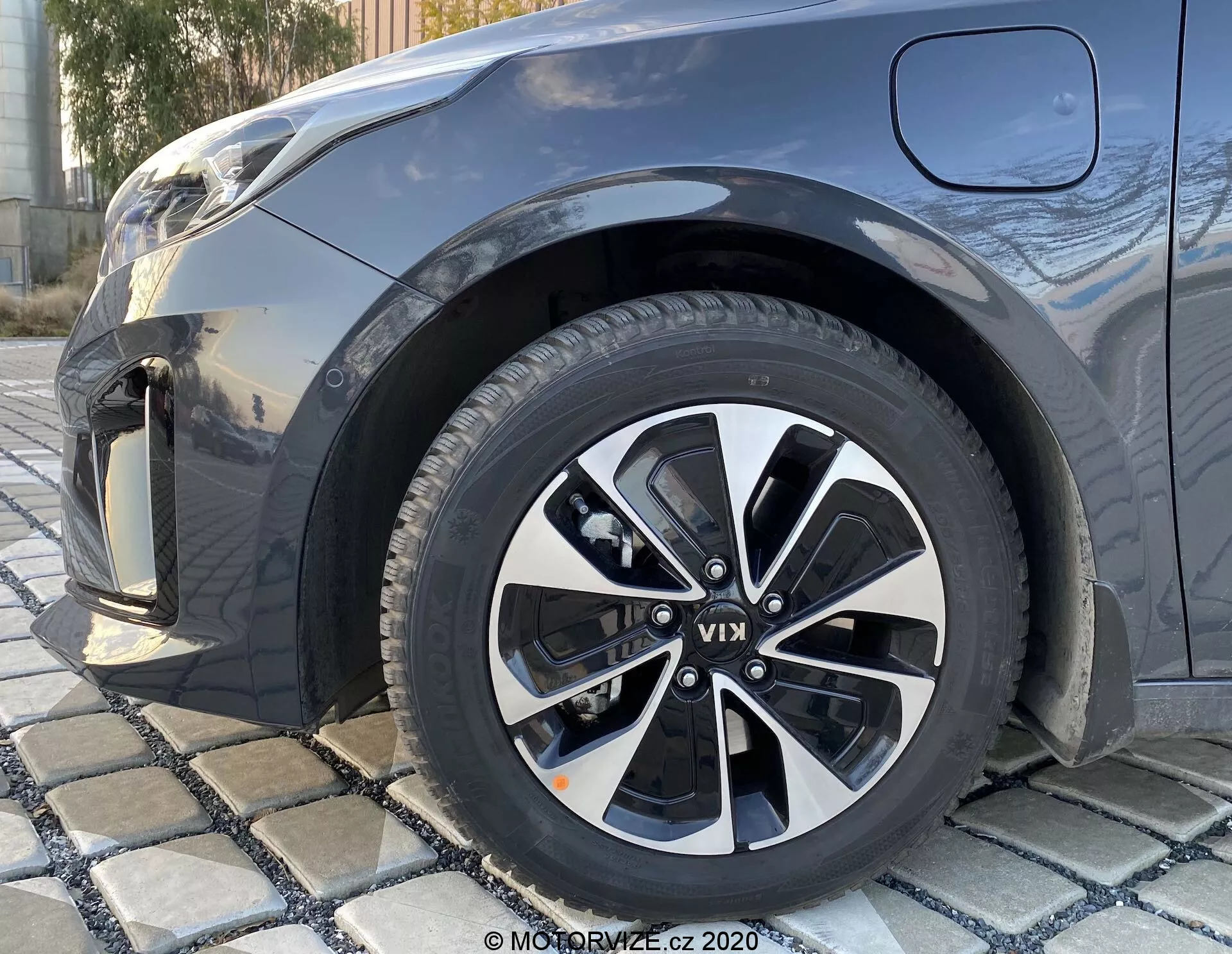Close-up em ângulo baixo da roda dianteira direita de um KIA Ceed 2018, com design de liga de raios múltiplos em preto e prata, com padrões de piso visíveis, mostrando parcialmente o para-lamas dianteiro, para-choque e parte do capô em uma cor metálica escura.