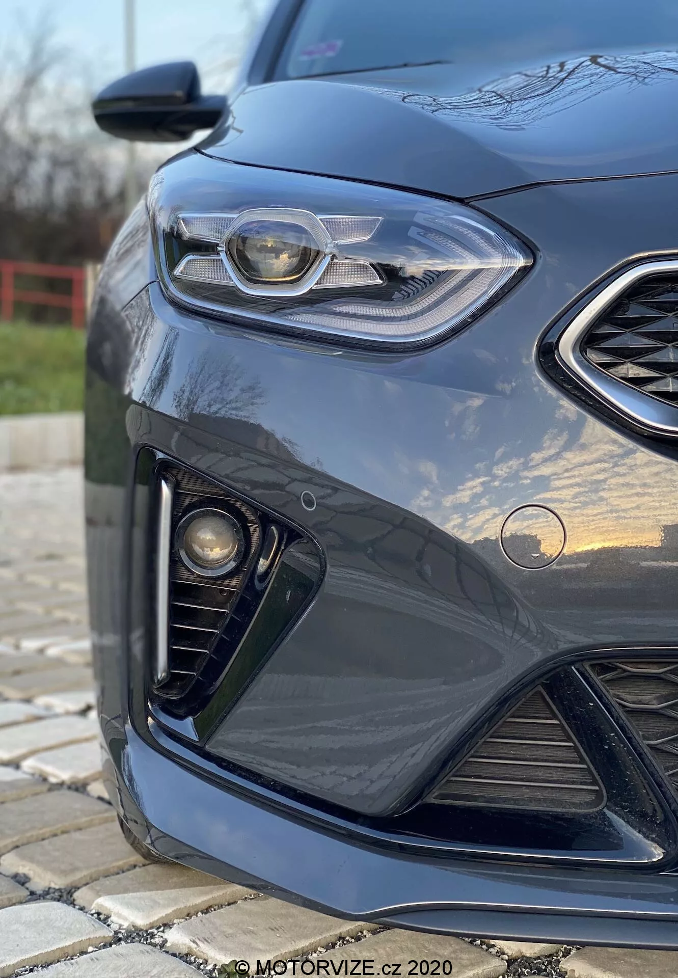 Vista aproximada e angular do lado frontal esquerdo de um Kia Ceed 2018 de cor escura, destacando o design elegante dos faróis com luz diurna em LED, grelha em favo de mel, conjunto de luzes de nevoeiro e para-choques dianteiro aerodinâmico, com fundo desfocado e reflexos na pintura brilhante.