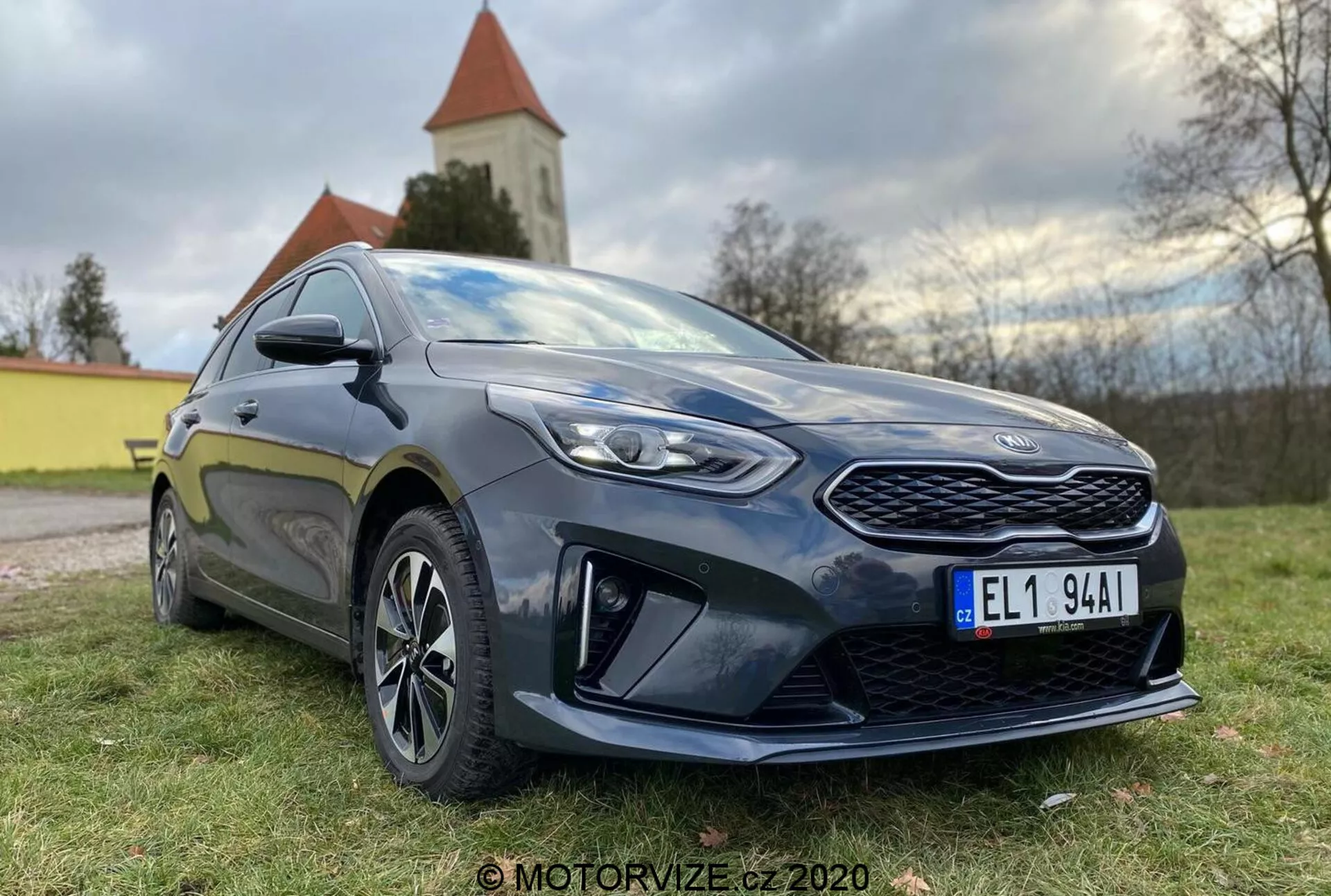 Vista detalhada de um moderno Kia Ceed 2018 hatchback fotografado de um ângulo baixo de três quartos frontais, mostrando uma grade proeminente, faróis de LED angulares e o perfil lateral até a porta do motorista. O carro é de cor escura, provavelmente azul escuro ou preto, com um brilho que destaca seu estado bem conservado. Os principais elementos de design incluem uma grade dianteira padronizada, faróis nítidos com lentes claras, elementos aerodinâmicos proeminentes no para-choque dianteiro inferior, rodas de liga leve de múltiplos raios, espelhos laterais da cor da carroceria com indicadores integrados e uma saia lateral esportiva. A postura do carro sugere um equilíbrio entre conforto e esportividade. A luz natural do dia com iluminação suave realça os detalhes, e ao fundo é visível uma paisagem natural com uma igreja ou torre.