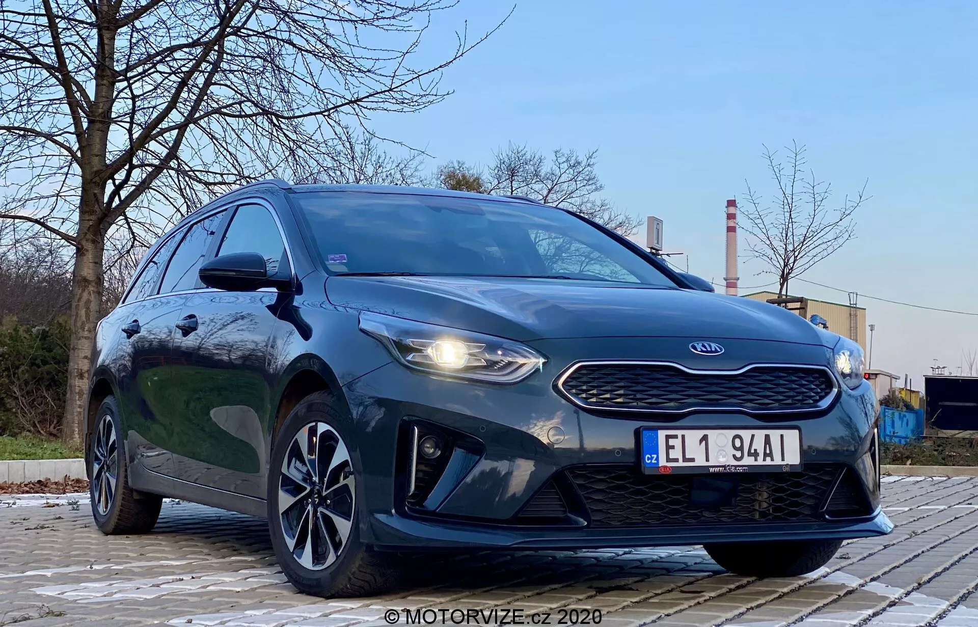 Fotografia do Kia Ceed 2018 tirada de um ângulo baixo e dianteiro esquerdo, enfatizando a grade, os faróis e o design da frente do veículo. O estilo agressivo do carro é destacado por faróis proeminentes, uma grade em forma de nariz de tigre e contornos no capô. A imagem também mostra luzes diurnas de LED, para-choque dianteiro aerodinâmico e rodas de liga leve elegantes. O ambiente é ao ar livre, com árvores e estruturas industriais ao fundo, proporcionando contraste. A pintura metálica escura do veículo é brilhante e bem iluminada, com reflexos indicando uma superfície limpa. A imagem foca no carro com uma perspectiva dramática de baixo ângulo sob iluminação externa suave.