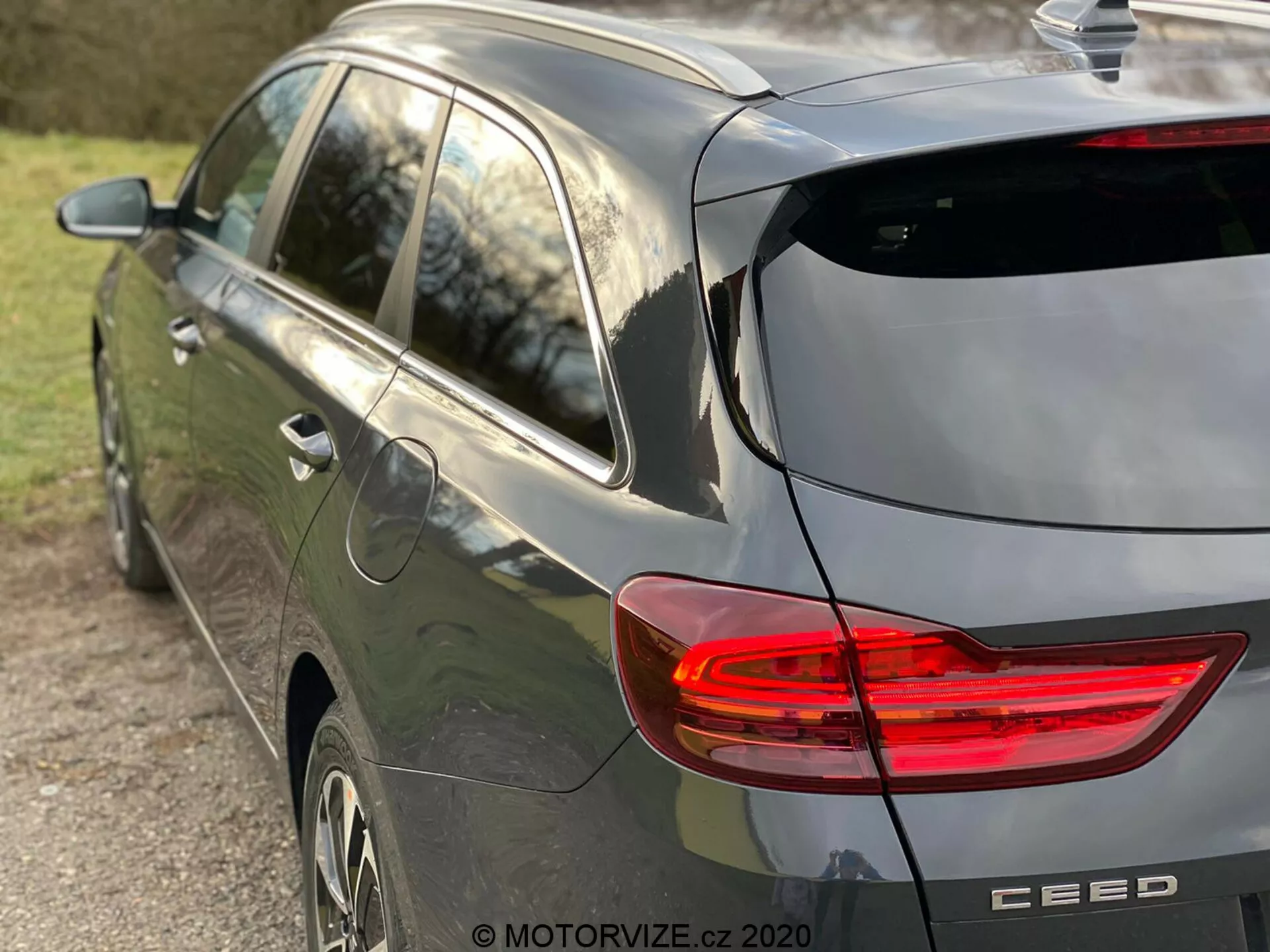 Vista parcial lateral e traseira de um carro hatchback Kia Ceed 2018 moderno de cor cinza, enfatizando a luz traseira LED iluminada em forma de C, design hatchback com limpador de para-brisa traseiro e discreto spoiler de teto, antena em forma de barbatana de tubarão, rodas de liga e emblema da marca com o nome do modelo 'CEED', capturado com luz natural de final de tarde e uma profundidade de campo rasa focando no veículo.
