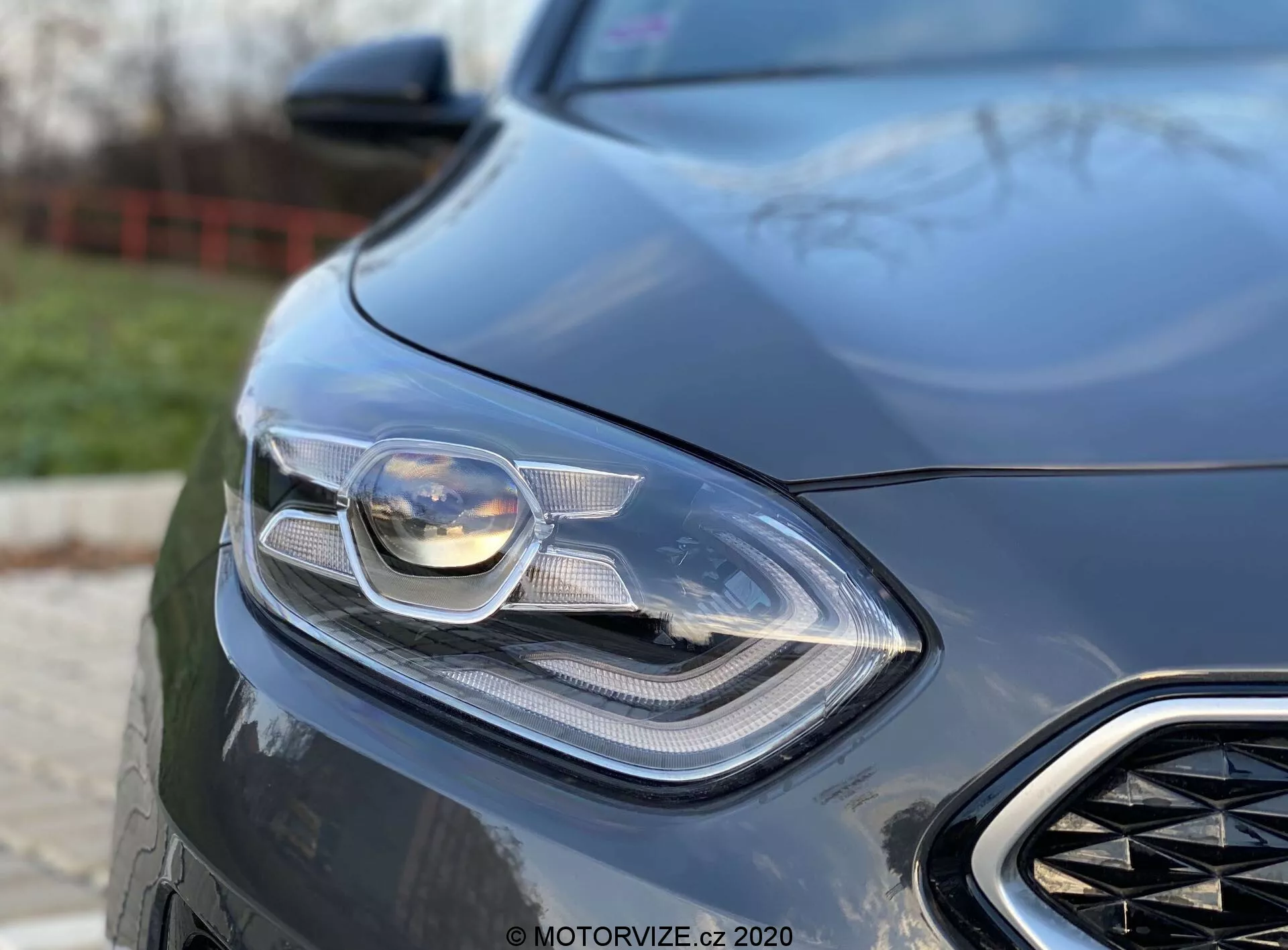 Close-up detalhado da porção dianteira esquerda de um Kia Ceed 2018, focando no seu farol sofisticado e parte da grade frontal. A foto é tirada de um ângulo baixo, destacando o design elegante e moderno do carro, com tecnologia de iluminação avançada visível, possivelmente incluindo faróis LED e adaptativos. O fundo desfocado acentua os elementos aerodinâmicos e a sensação de luxo do carro, indicando um veículo orientado para desempenho.