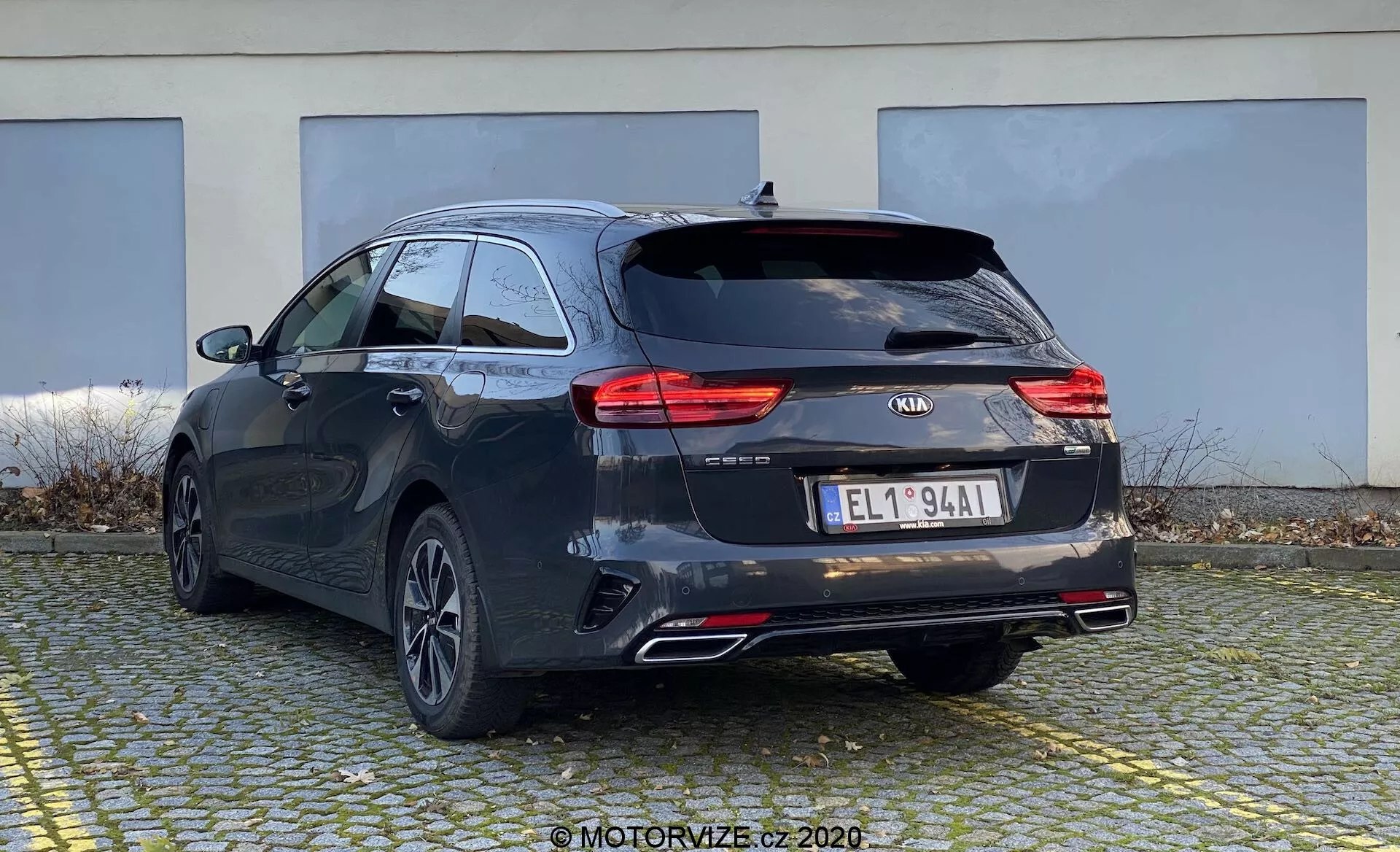 A imagem mostra um Kia Ceed 2018 em uma perspectiva traseira de três quartos, destacando a traseira e a lateral do carro station wagon. O carro exibe um tom escuro, possivelmente preto ou cinza escuro, com linhas características e um teto ligeiramente inclinado para um visual esportivo. Visíveis estão as luzes traseiras de LED modernas, o logotipo da Kia, o nome do modelo 'CEED', emblemas adicionais, uma placa de licença estilo europeu, rodas traseiras multiraios de liga leve, tubos de escape duplos e um limpador traseiro. Sugere-se, mas não visível, uma câmera de ré e sensores de estacionamento. O pequeno spoiler de teto e os trilhos do teto indicam opções adicionais de carga. O carro está estacionado em uma área pavimentada com um prédio ao fundo, mostrando a parte traseira e parte do perfil lateral de um ângulo ligeiramente baixo.