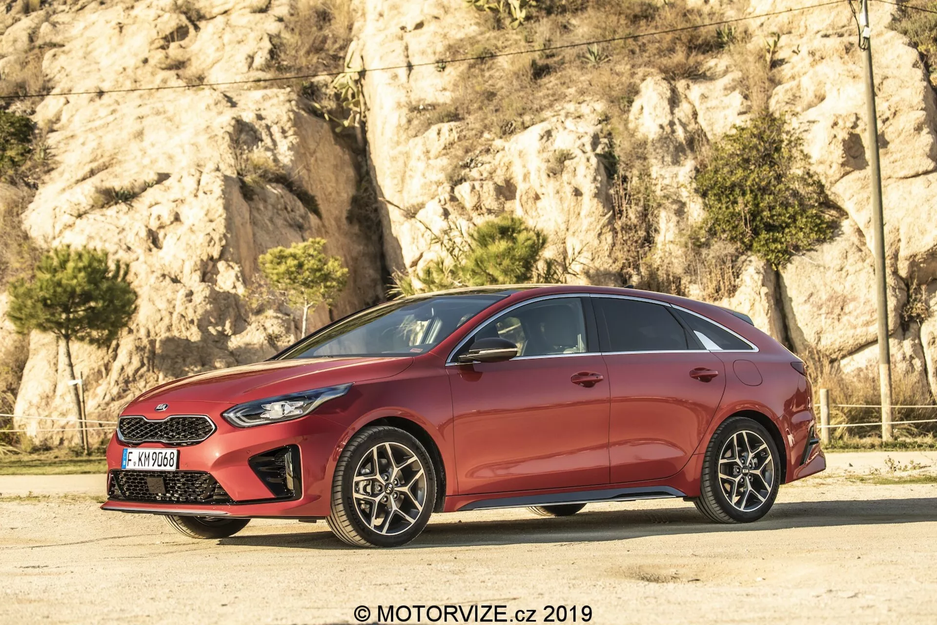 Vista de perfil lateral de um hatchback Kia Ceed vermelho 2021, com um design elegante e aerodinâmico, linhas de teto inclinadas e linhas contornadas nas portas. O carro, um modelo de cinco portas, tem acabamento cromado nas janelas e pilares B escurecidos, criando um efeito de painel de vidro contínuo. As rodas de liga leve de múltiplos raios, com freios a disco visíveis, adicionam um toque esportivo. Faróis afiados e a grade parcialmente visível marcam a frente, enquanto os espelhos laterais na cor da carroceria com sinais de giro integrados melhoram a visibilidade e a segurança.
