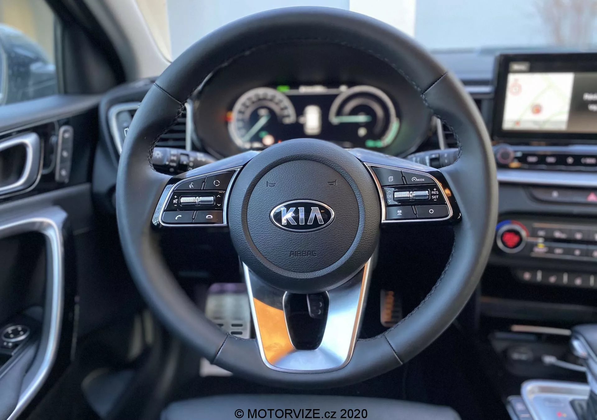 Foto do interior de um Kia Ceed 2018 focando no volante e no painel de instrumentos visto da perspectiva do motorista. O volante moderno possui controles integrados para controle de cruzeiro, interação multimídia e comunicação viva-voz, com o logo da Kia em destaque. Atrás do volante está o painel de instrumentos, que combina mostradores analógicos e um display digital mostrando o velocímetro e o tacômetro, junto com um display multifuncional. À direita do volante, parte do console central é visível, contendo um sistema de infoentretenimento com tela sensível ao toque e navegação GPS, além dos controles de climatização. A imagem é clara, com profundidade de campo rasa que destaca o volante e seus arredores imediatos.