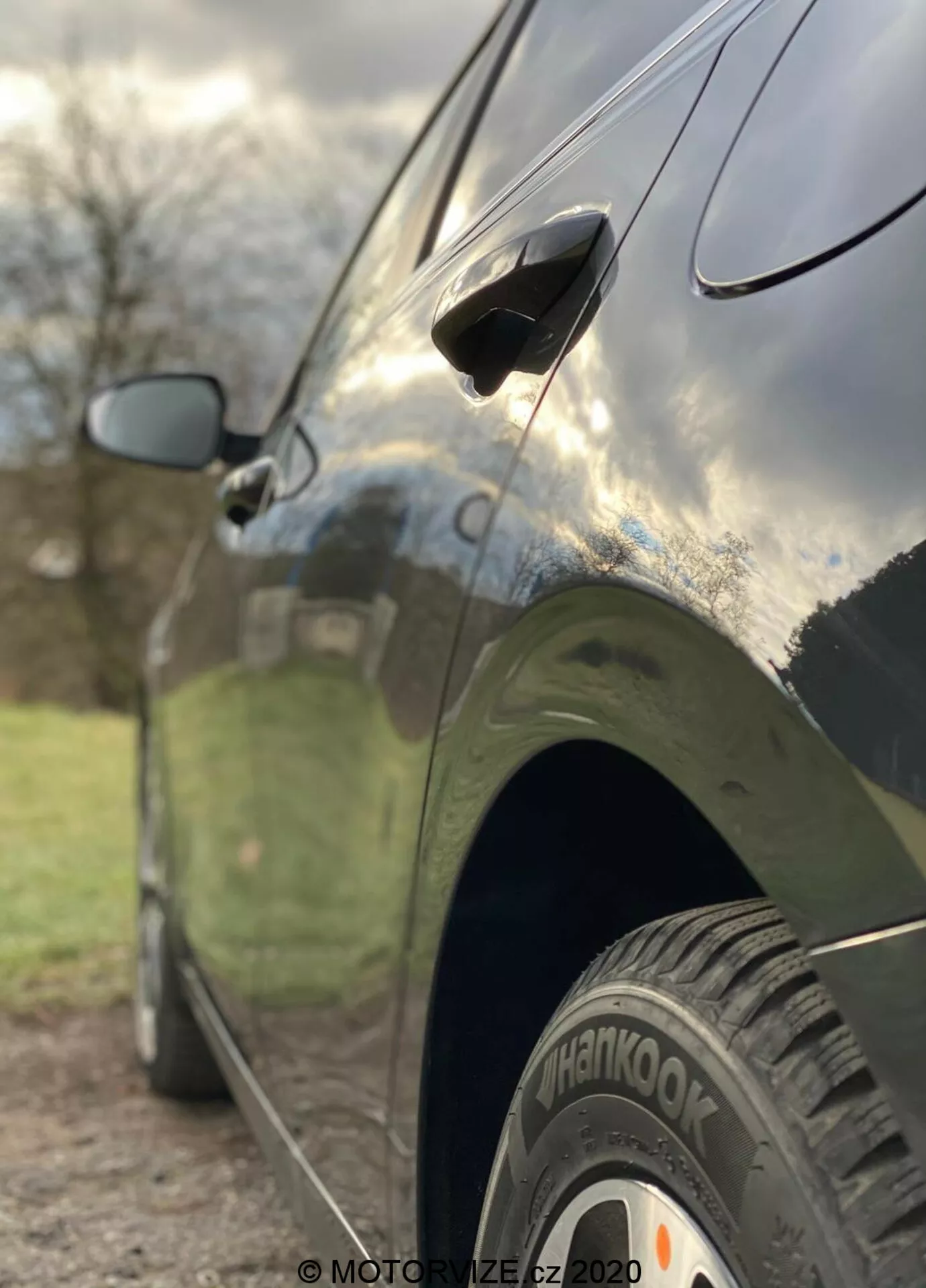 Vista lateral em close-up do Kia Ceed 2018, destacando características como a roda traseira com pneu 'Hankook', maçaneta de porta aerodinâmica e espelho lateral parcialmente desfocado. A foto é tirada de um ângulo baixo com uma profundidade de campo reduzida que coloca nítidamente o pneu traseiro e a parte inferior da porta do carro em foco, enquanto o fundo é artisticamente desfocado. O carro tem um acabamento escuro e brilhante que reflete o céu e os arredores, mostrando linhas de design elegantes e uma carroceria bem mantida.