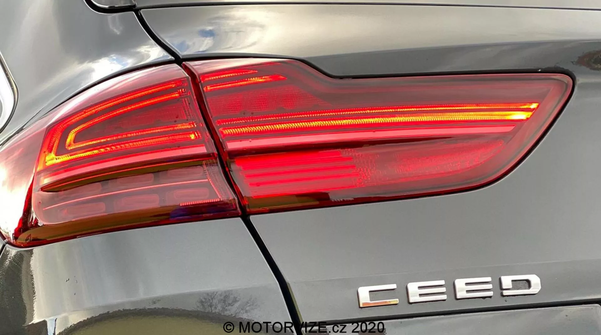 Vista detalhada e aproximada do conjunto de luz traseira direita do Kia Ceed 2018, iluminada, exibindo um design moderno com linhas nítidas e angulares, seções de lente clara e vermelha, e distintivo do modelo 'CEED'. Linha horizontal separa a luz traseira do para-choque traseiro. Reflexos na carroceria destacam a curvatura da superfície. Marca d'água 'MOTOR1.cz ©2020'.