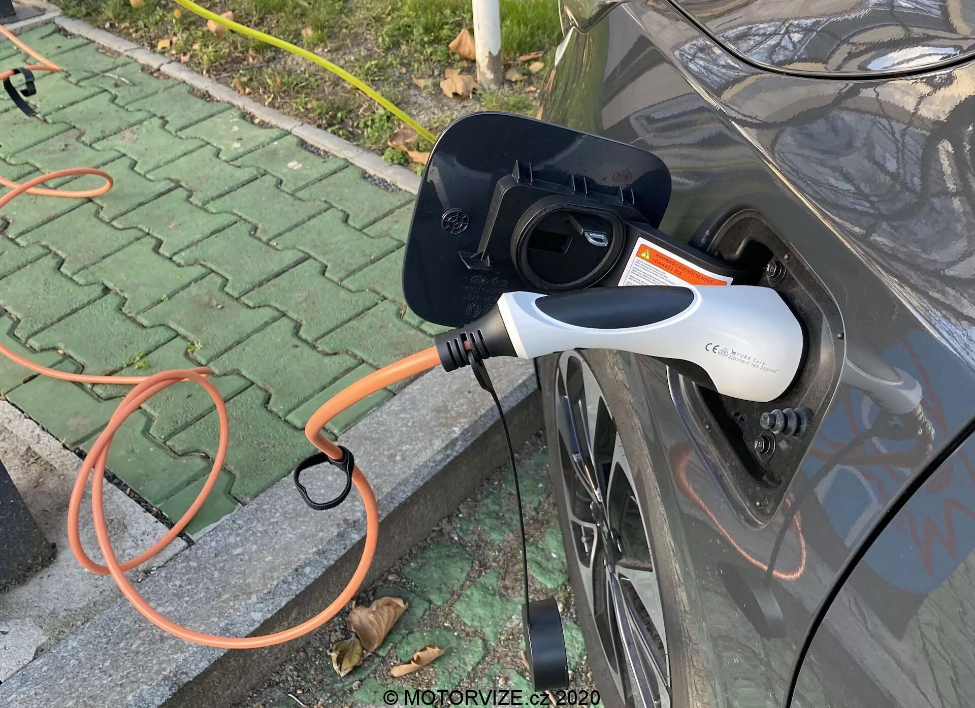 Vista lateral em close-up de um veículo elétrico (EV) cinza em carga, destacando o porto de carregamento aberto e um conector EVSE laranja conectado ao carro. O conector de carregamento moderno branco e preto possui uma luz indicadora LED. A imagem foca na interação de carregamento, situada em uma área pavimentada ao ar livre com folhas espalhadas.