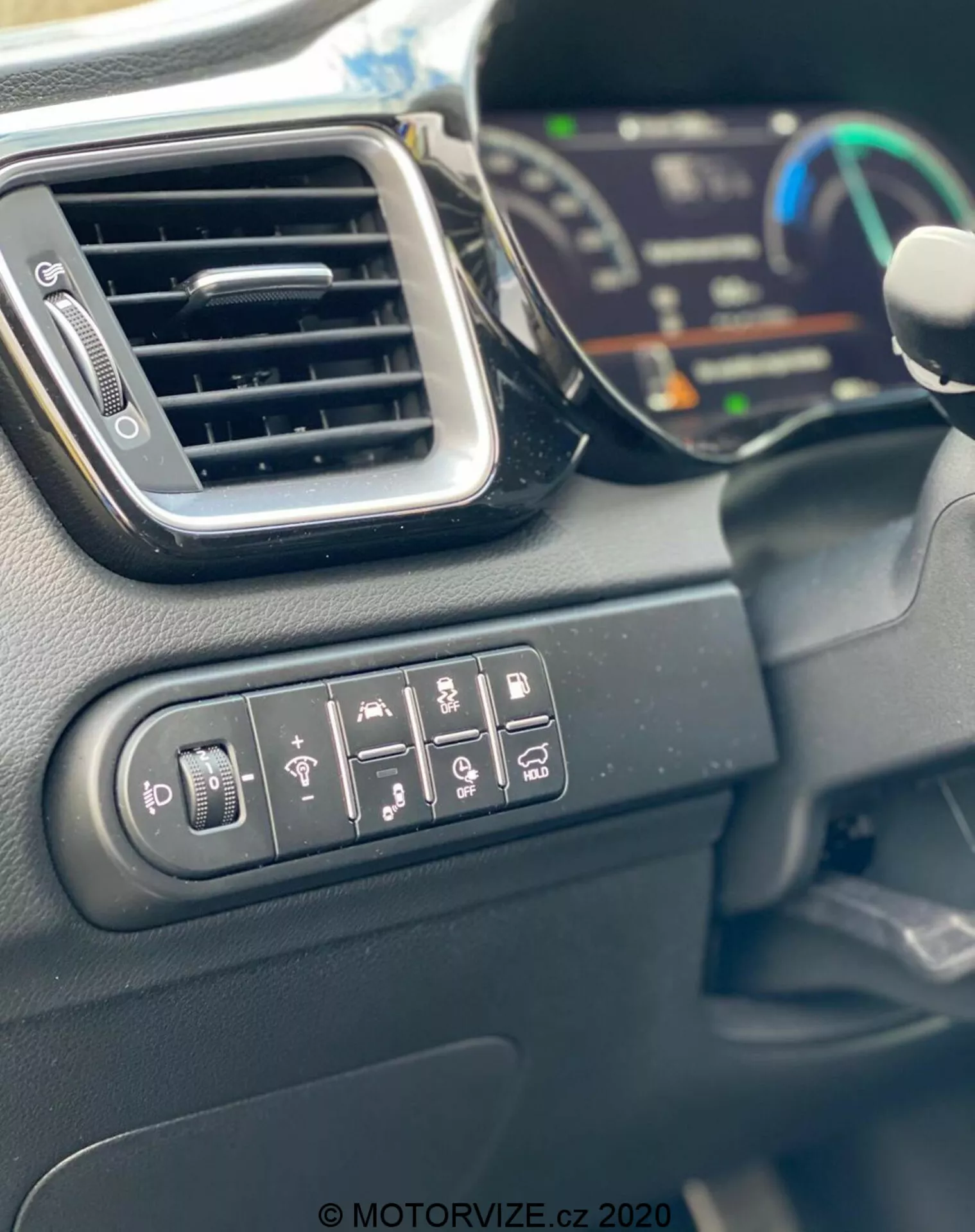Vista de perto do interior do Kia Ceed 2018 mostrando os botões de controle para ajuste dos espelhos laterais, aquecimento da janela traseira, aquecimento do volante, aviso de saída de faixa, assistência de estacionamento e controle de tração. A imagem também apresenta uma ventilação de ar elegante, parte de um painel de instrumentos digital em segundo plano desfocado e a borda do volante. Os materiais internos são principalmente plásticos duros com acabamento fosco, capturados à luz do dia.