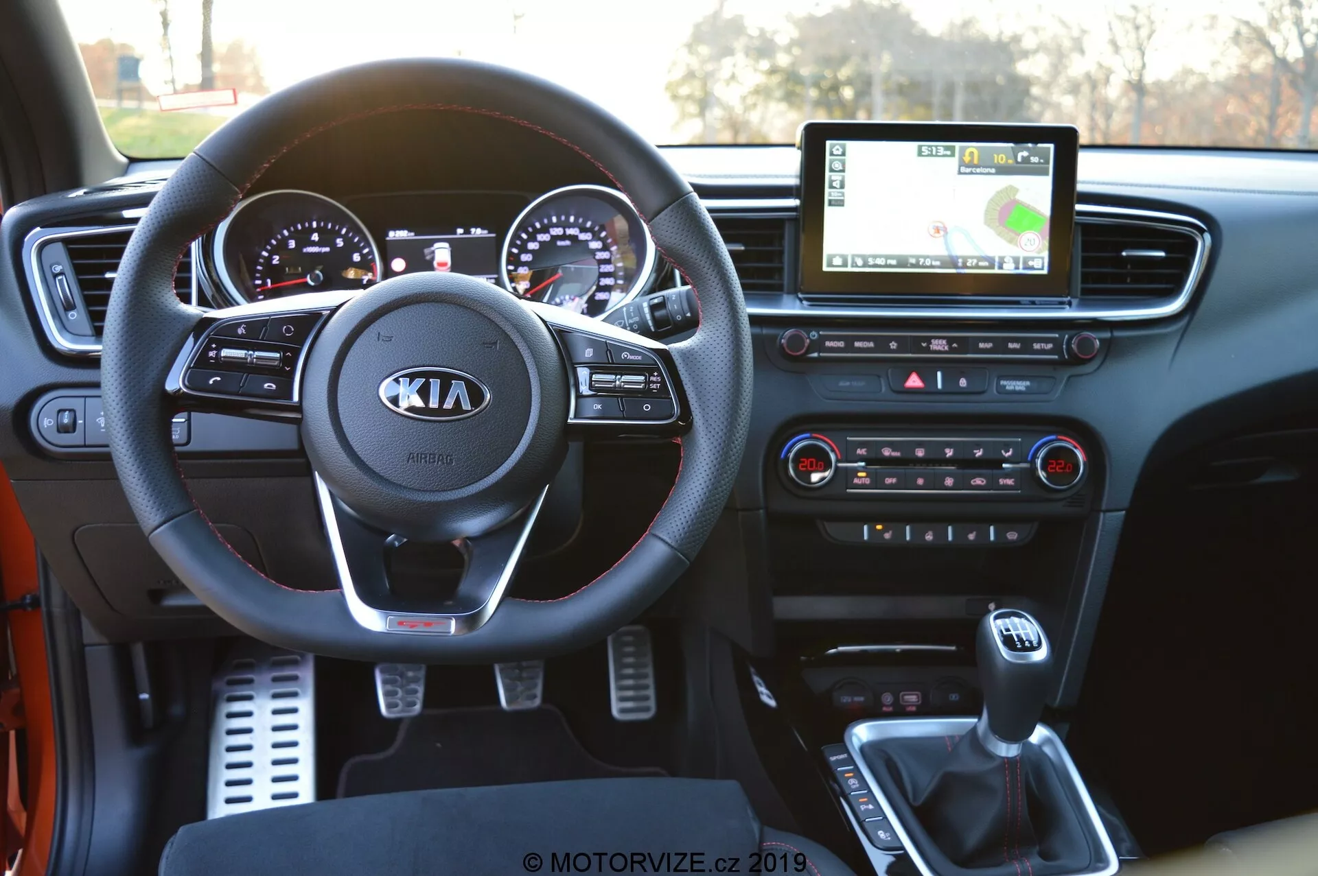 Vista interna de um Kia Ceed 2021 da perspectiva do motorista, mostrando o painel detalhado. A imagem apresenta de forma proeminente o volante com o logotipo da Kia e os controles incorporados para áudio, chamadas e controle de cruzeiro. Atrás dele, você pode ver o painel de instrumentos com medidores analógicos acentuados em cromo e um display digital. A tela central de infotainment mostra a navegação GPS, enquanto abaixo estão os botões e botões de controle do clima. A consola central mostra a alavanca de câmbio, porta-copos e uma porta USB para conectividade de dispositivos, destacando a tecnologia automotiva moderna.